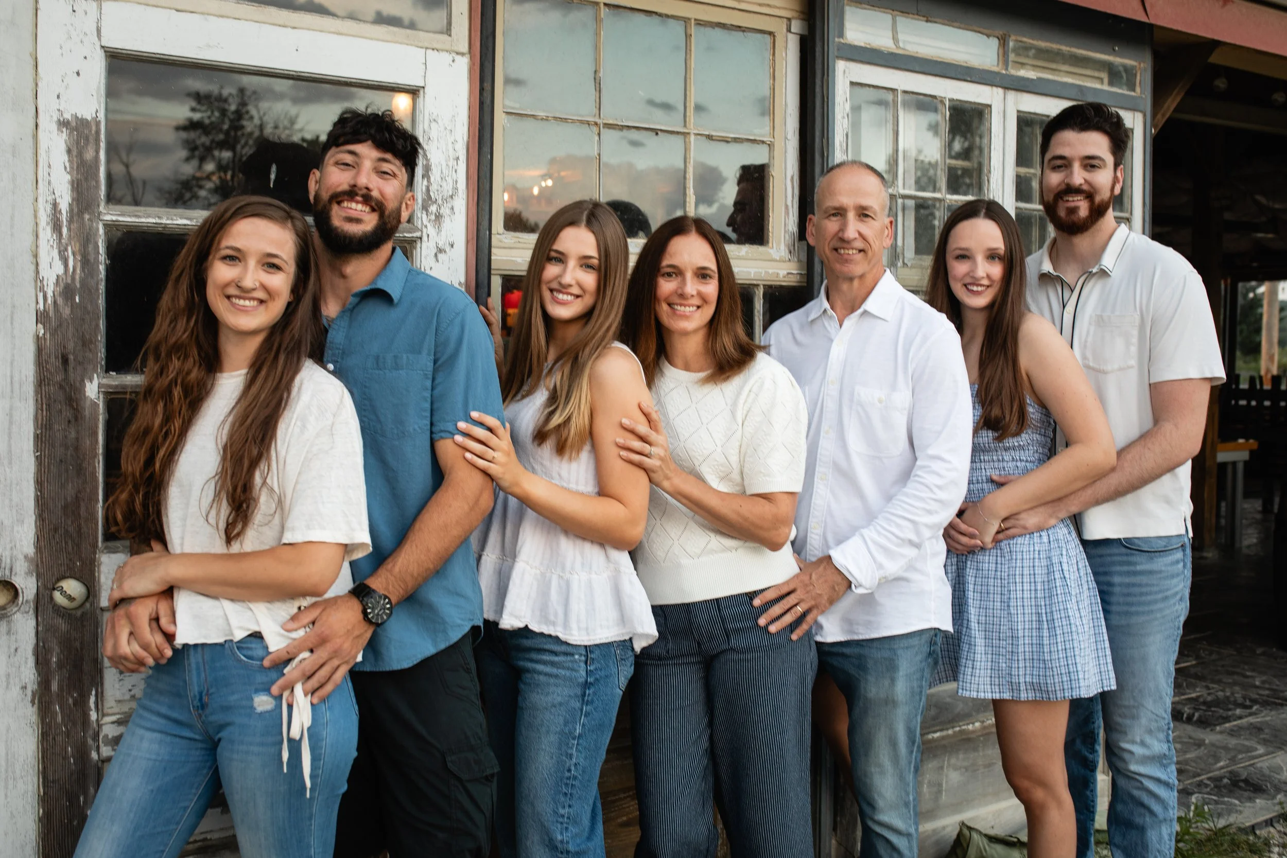 A group of seven people smiling and posing outside a rustic building with large windows, during daylight.
