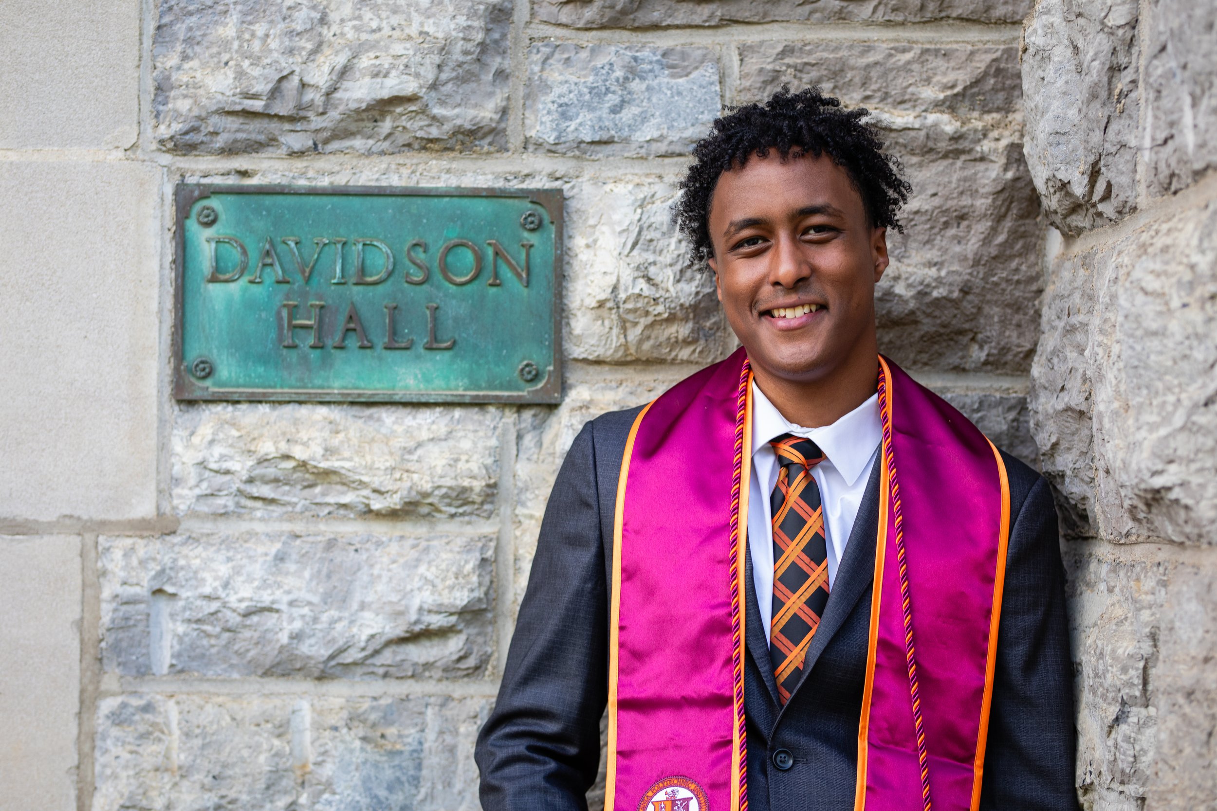 Young man in a suit with a graduation sash standing next to a stone wall with a green plaque that reads 'Davidson Hall'.