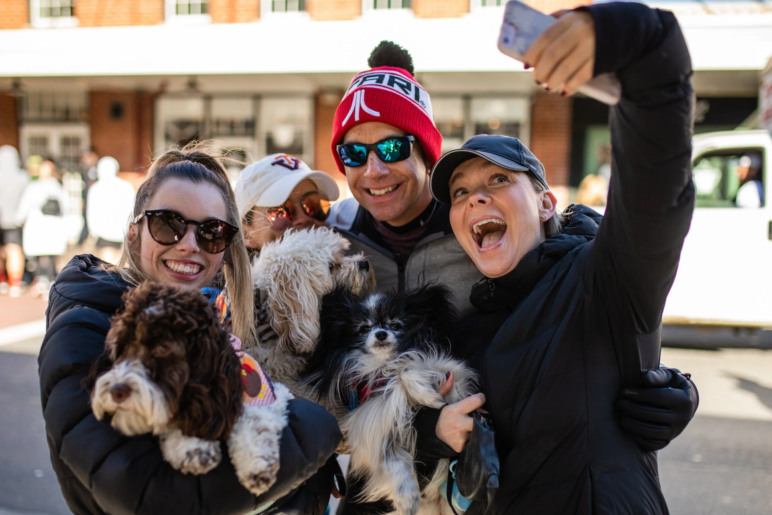 A group of people smiling and taking a selfie outdoors, holding small dogs in their arms.