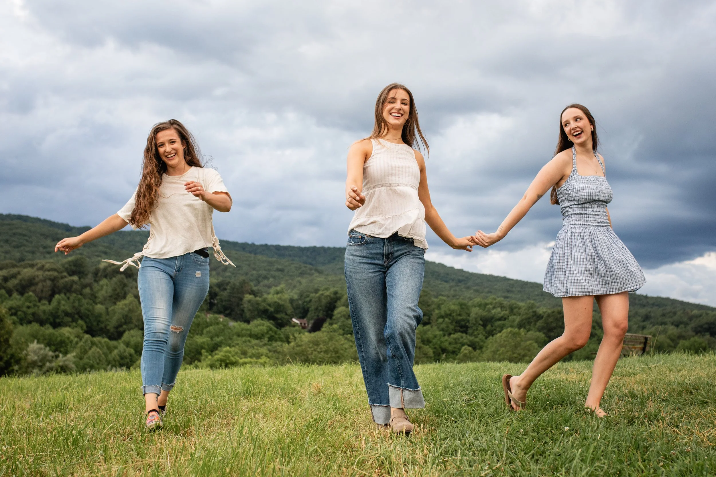 Three young women holding hands and smiling while walking on a grassy field with green hills and cloudy sky in the background.