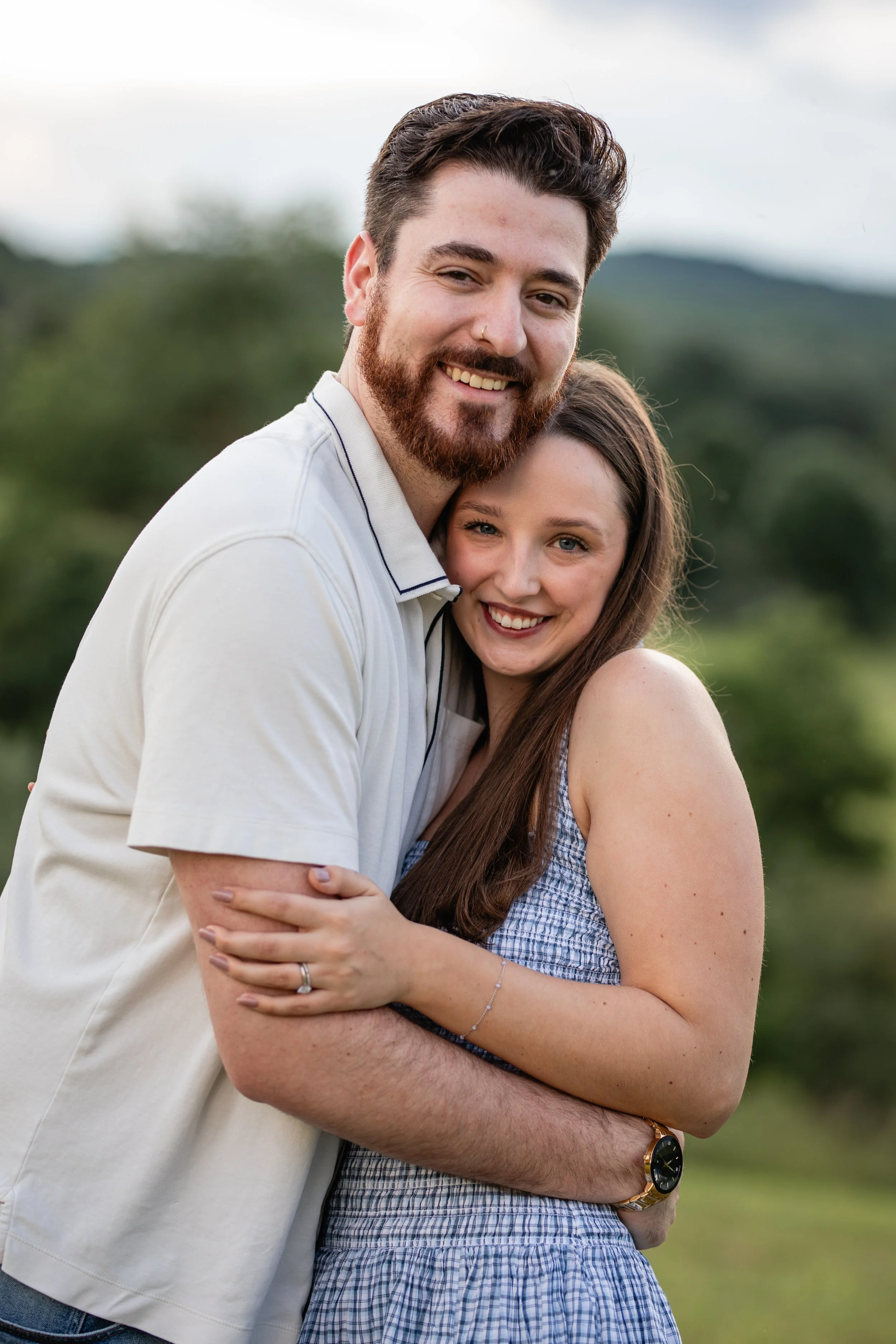 A happy couple embracing outdoors on a cloudy day, smiling at the camera with a green, mountainous landscape in the background.