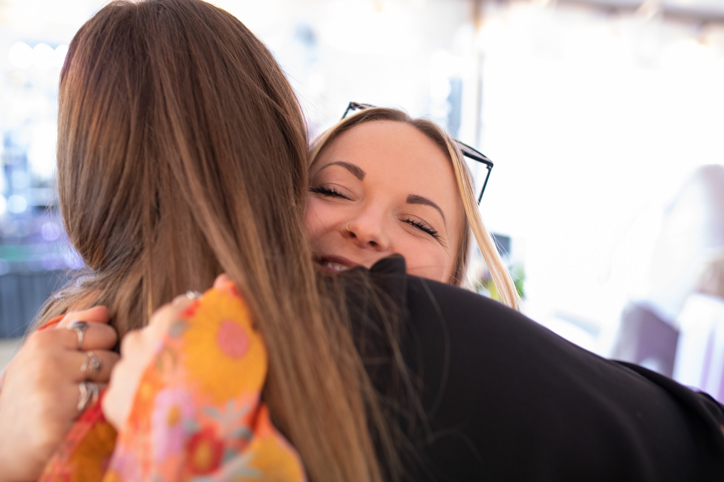 Two women hugging and smiling indoors, one woman with long brown hair and rings on her fingers, the other woman with blonde hair and glasses, wearing a floral dress.