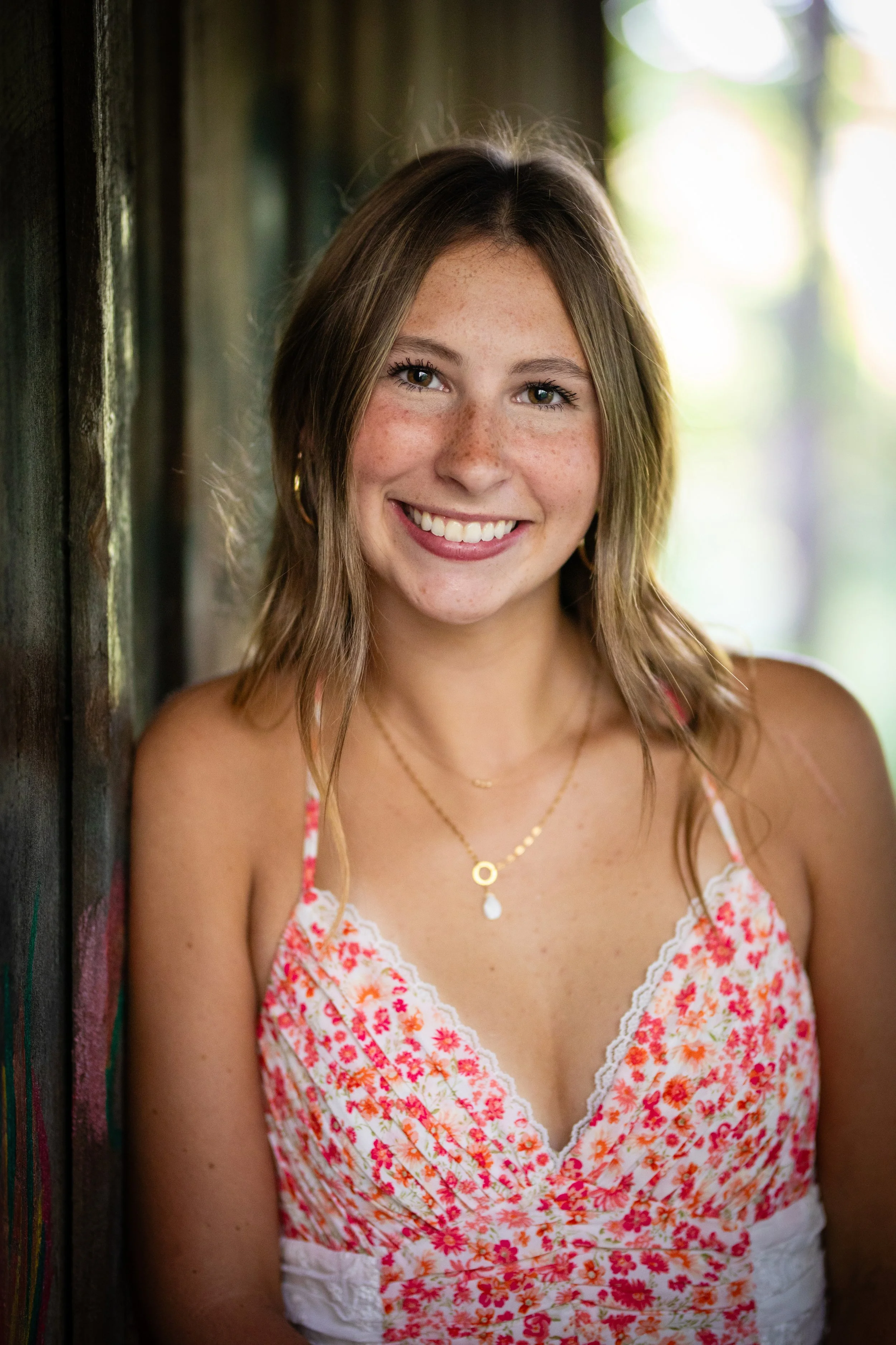 A young woman with light brown hair, freckles, and a bright smile, wearing a floral dress and layered necklaces, leaning against a wooden wall outdoors.