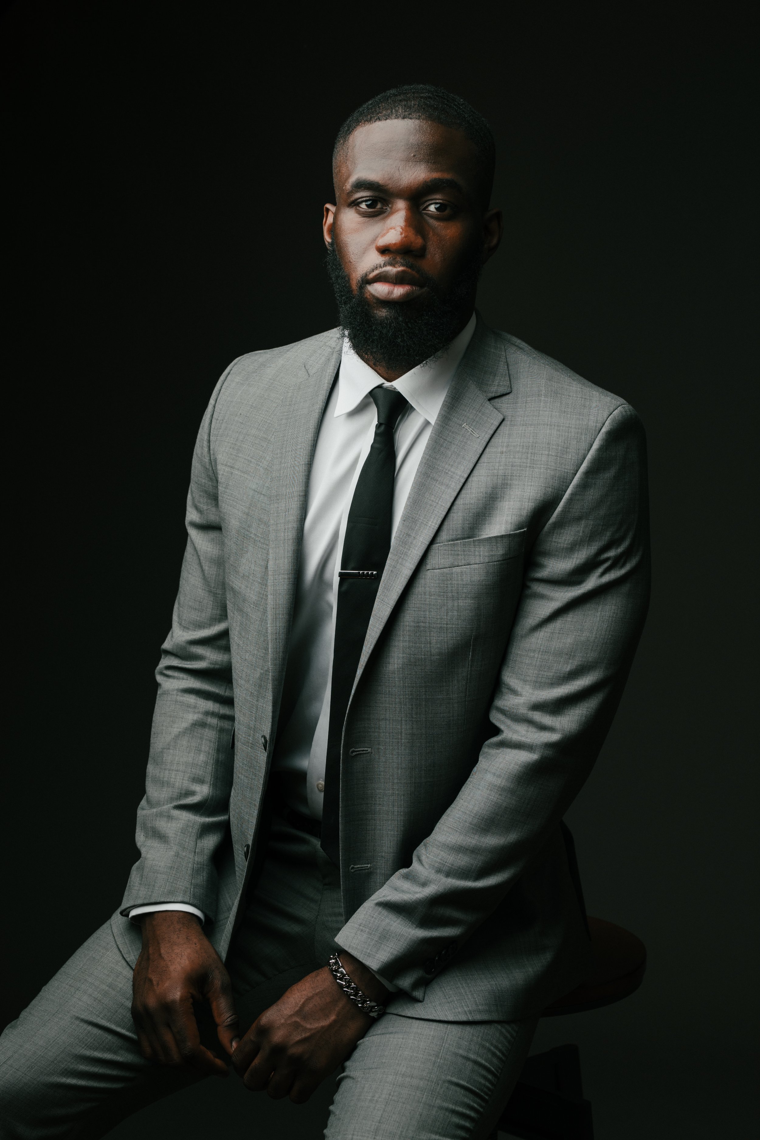 A man in a gray suit with a white shirt and black tie sitting on a stool against a black background.