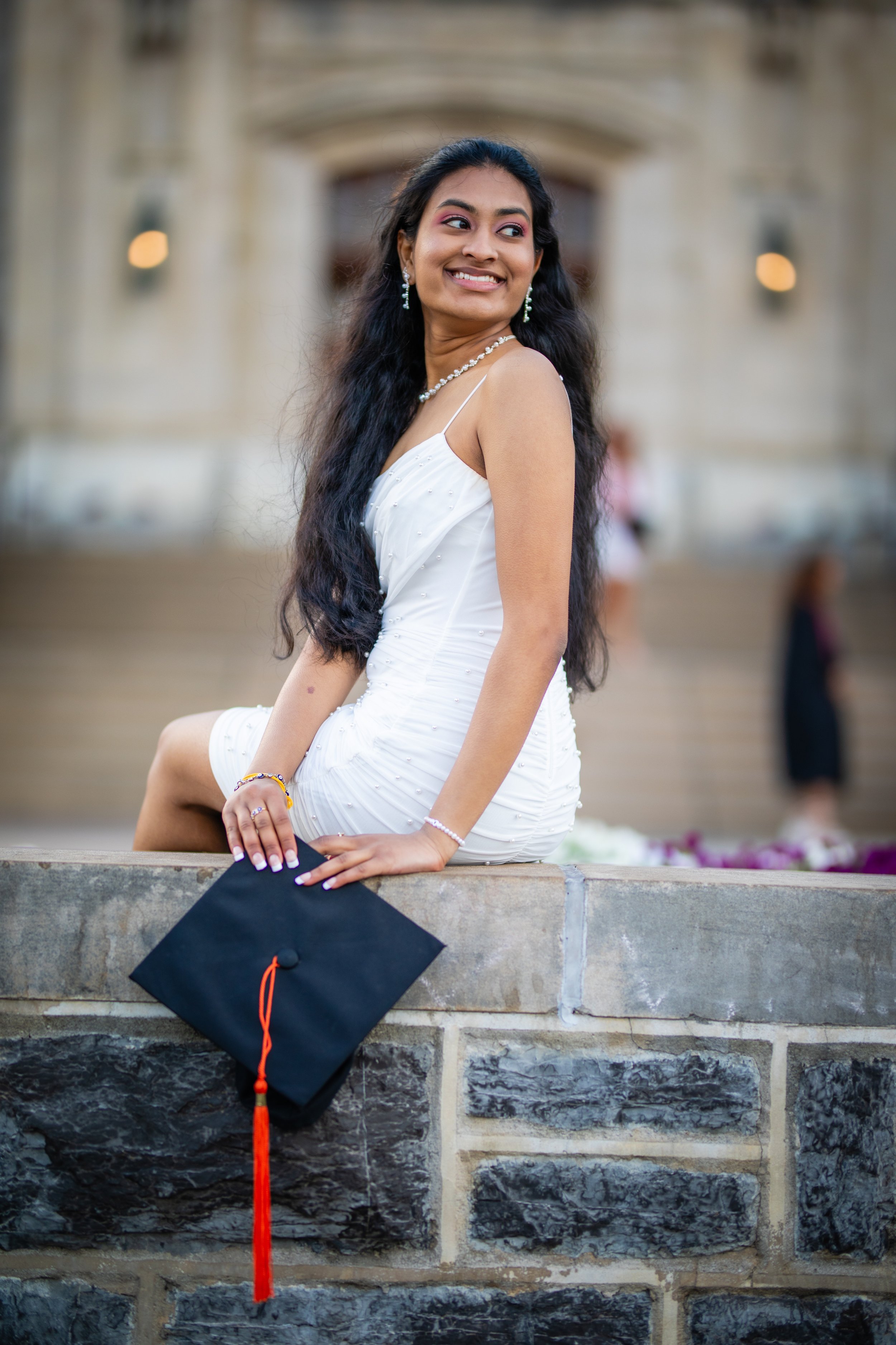A young woman in a white dress sitting on a stone wall, holding a graduation cap with a red tassel, smiling and looking to the side, with a historic building in the background.