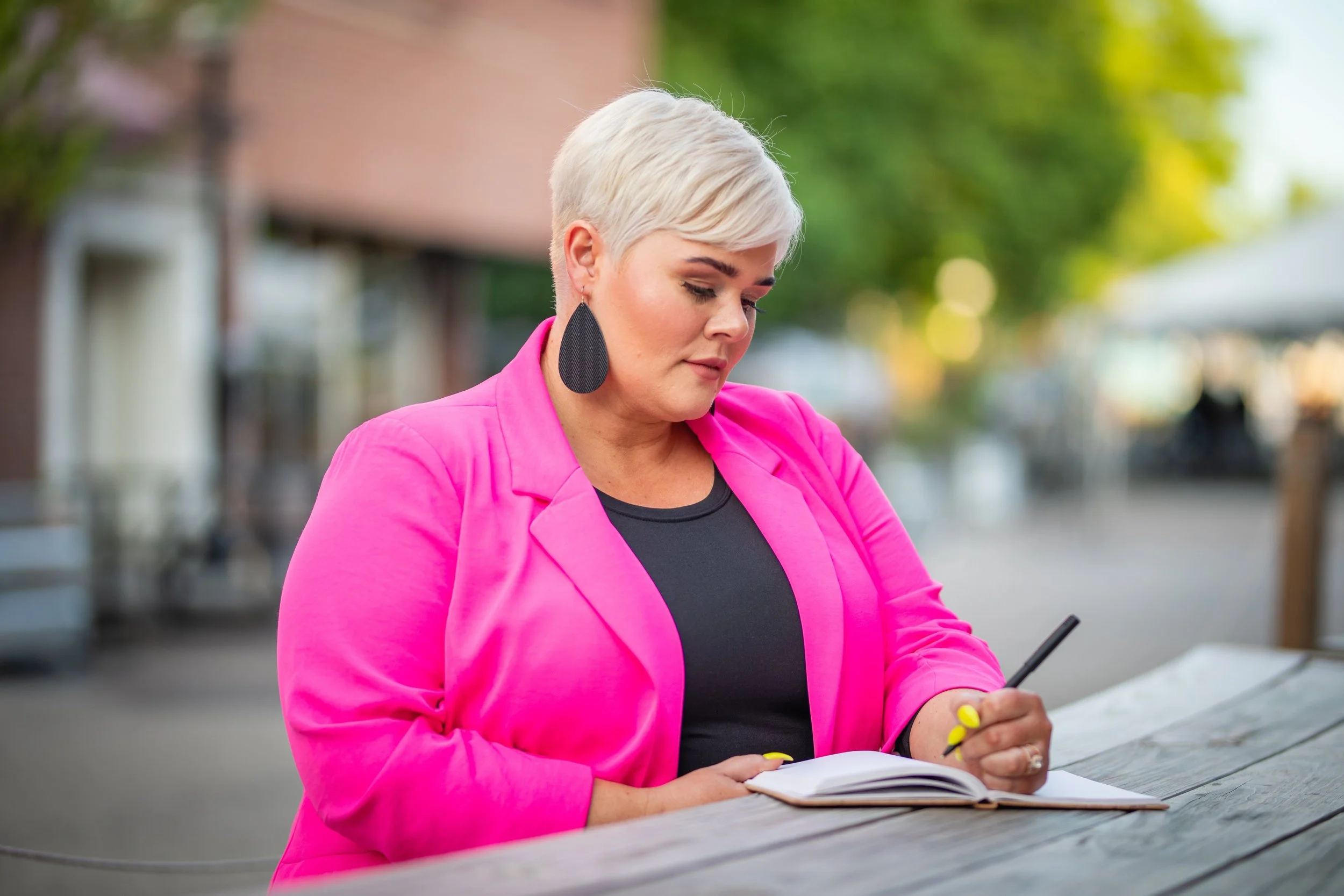 A woman with short blonde hair, wearing a bright pink blazer, sitting at an outdoor table, writing in a notebook with a pen, with a blurred background of trees and buildings.