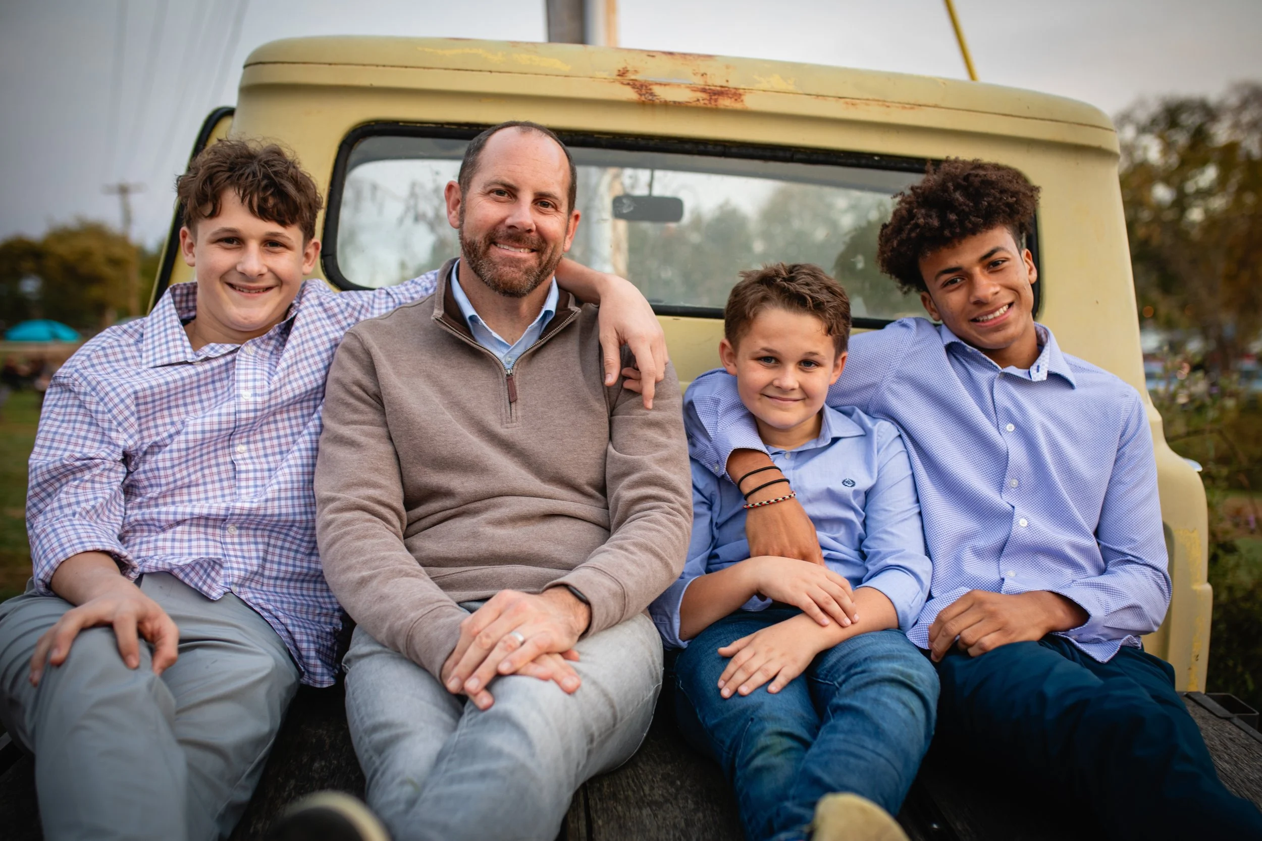 Five smiling people sitting together on the back of a yellow vehicle, including a man, two boys, and two young men, outdoors during the daytime.