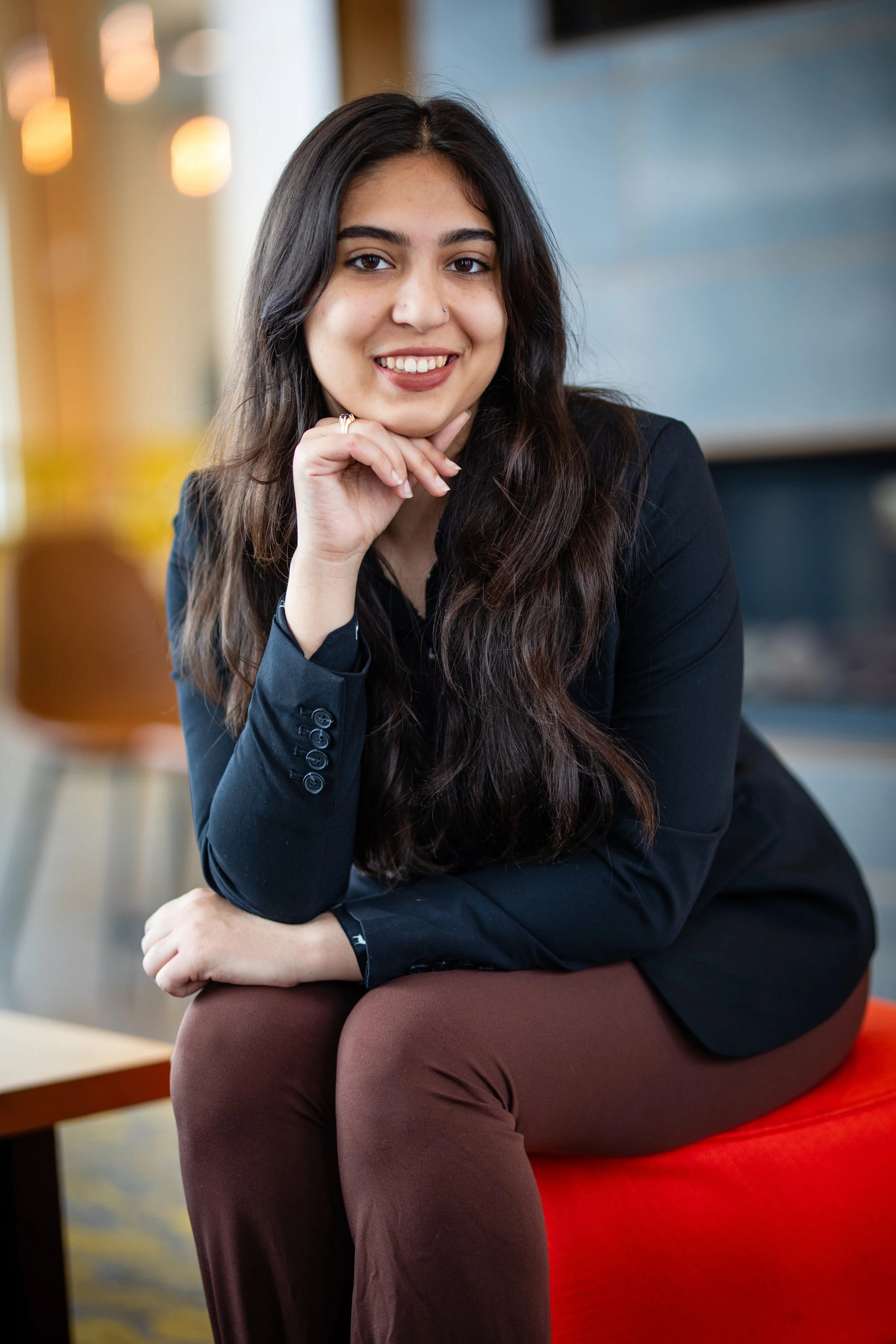 A young woman with long dark hair, wearing a black blazer and brown pants, sitting on a red chair in a modern, well-lit space, smiling with her chin resting on her hand.