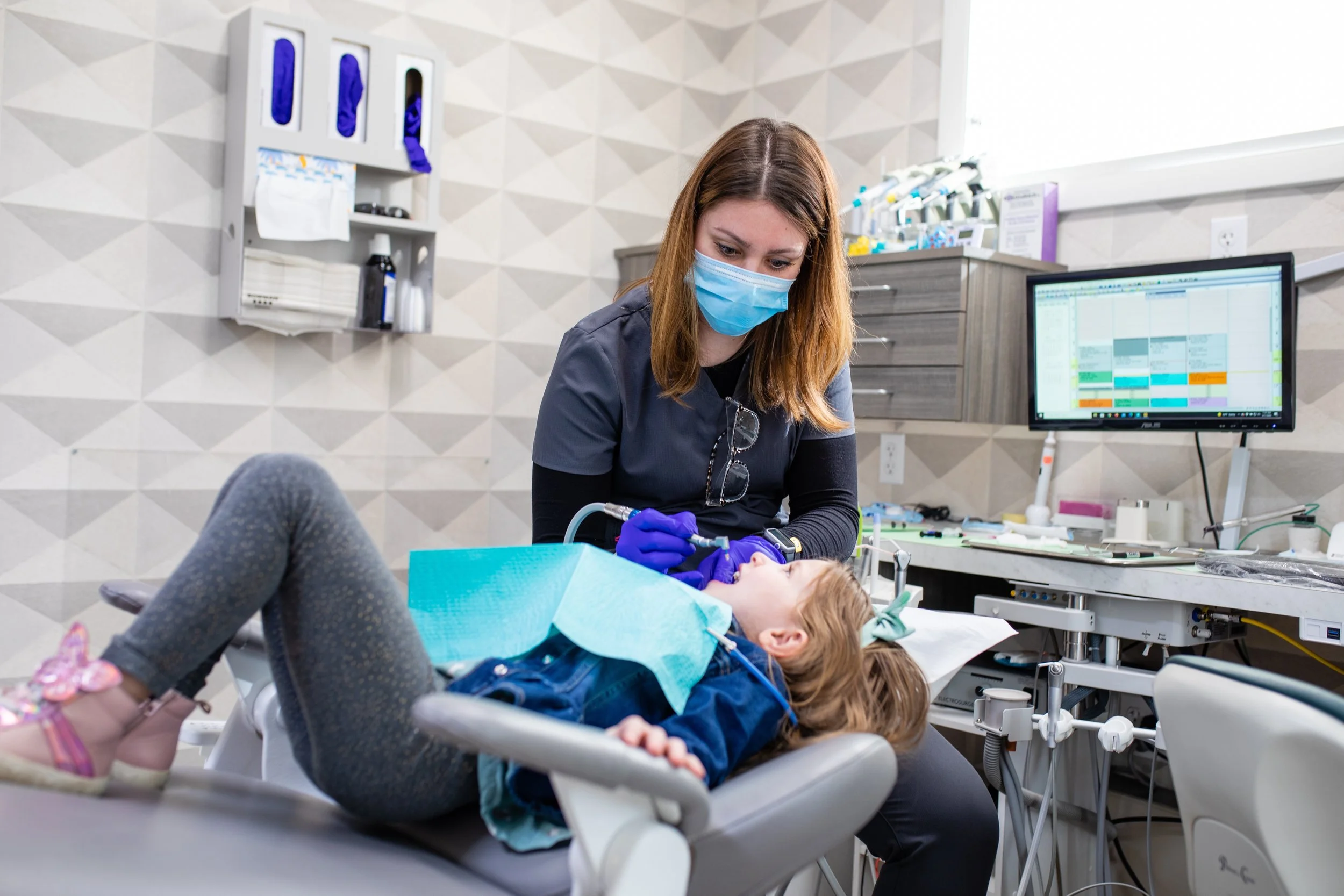 Dentist or dental hygienist performing a dental procedure on a young girl in a dental clinic, with dental tools, a computer screen, and dental supplies in the background.