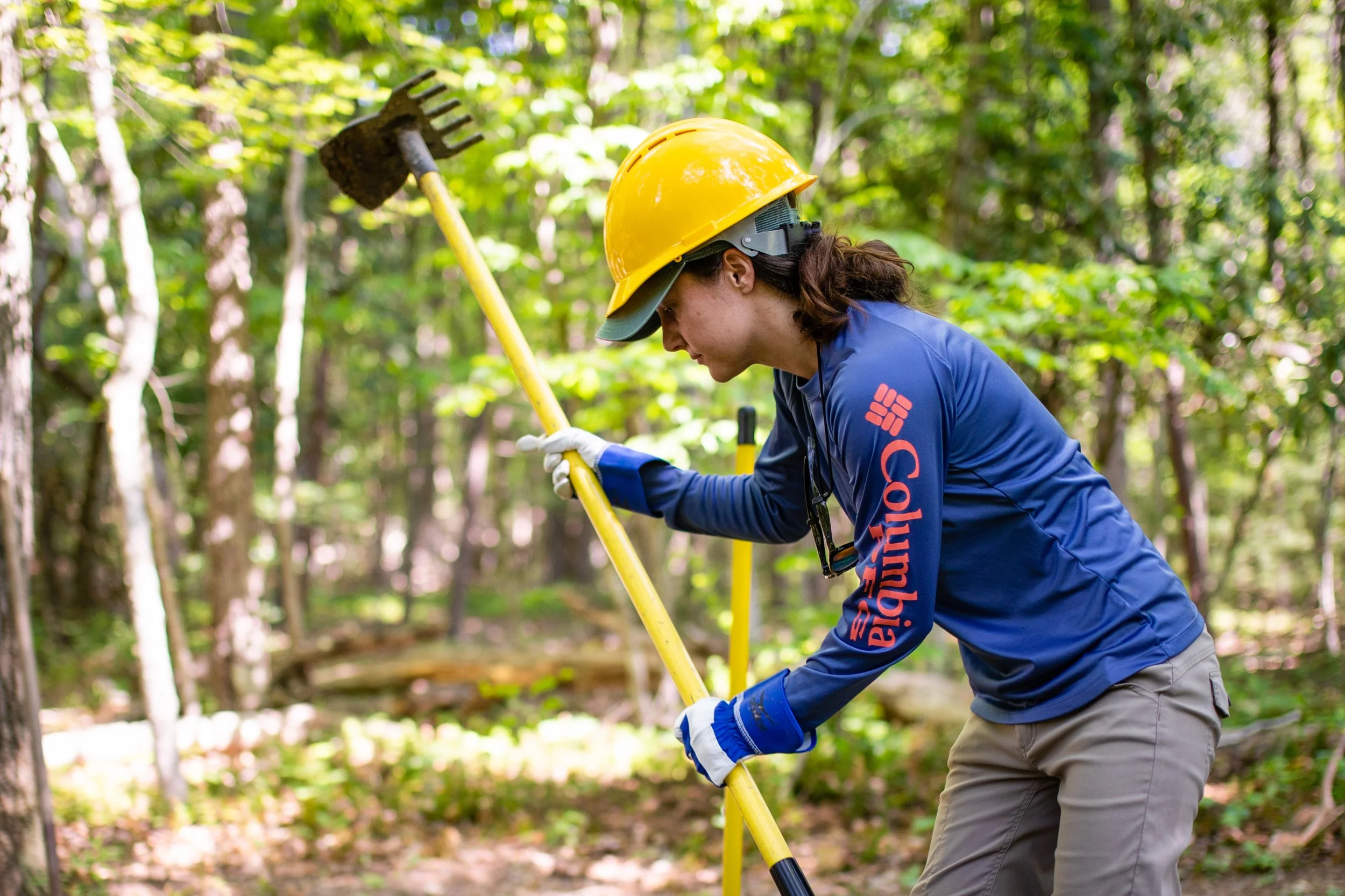 A female worker in a yellow safety helmet and blue Columbia jacket is using a survey tool in a forested area.