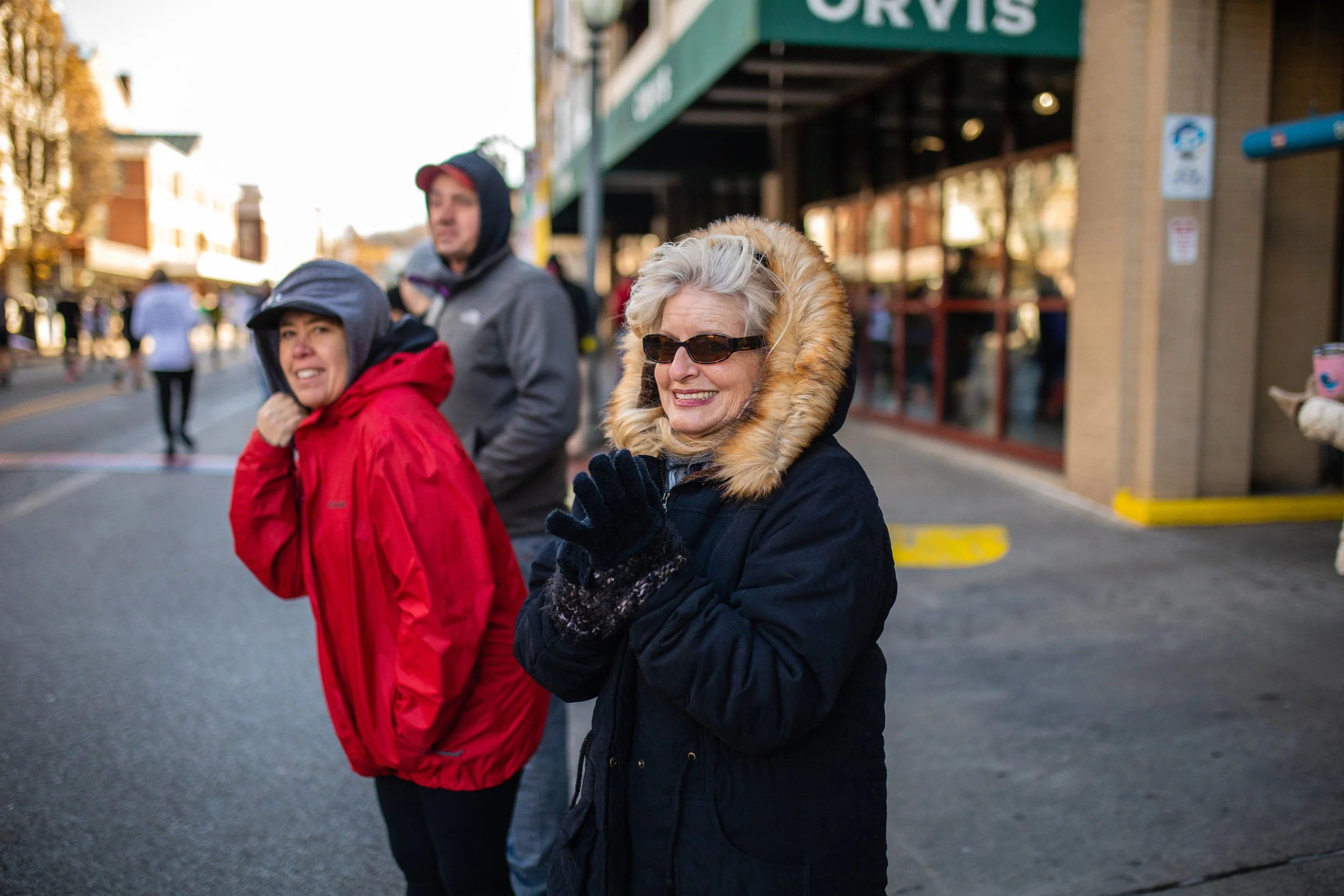 Three people standing on a city street during cold weather, wearing winter jackets and gloves, with a woman smiling and clapping in the foreground.