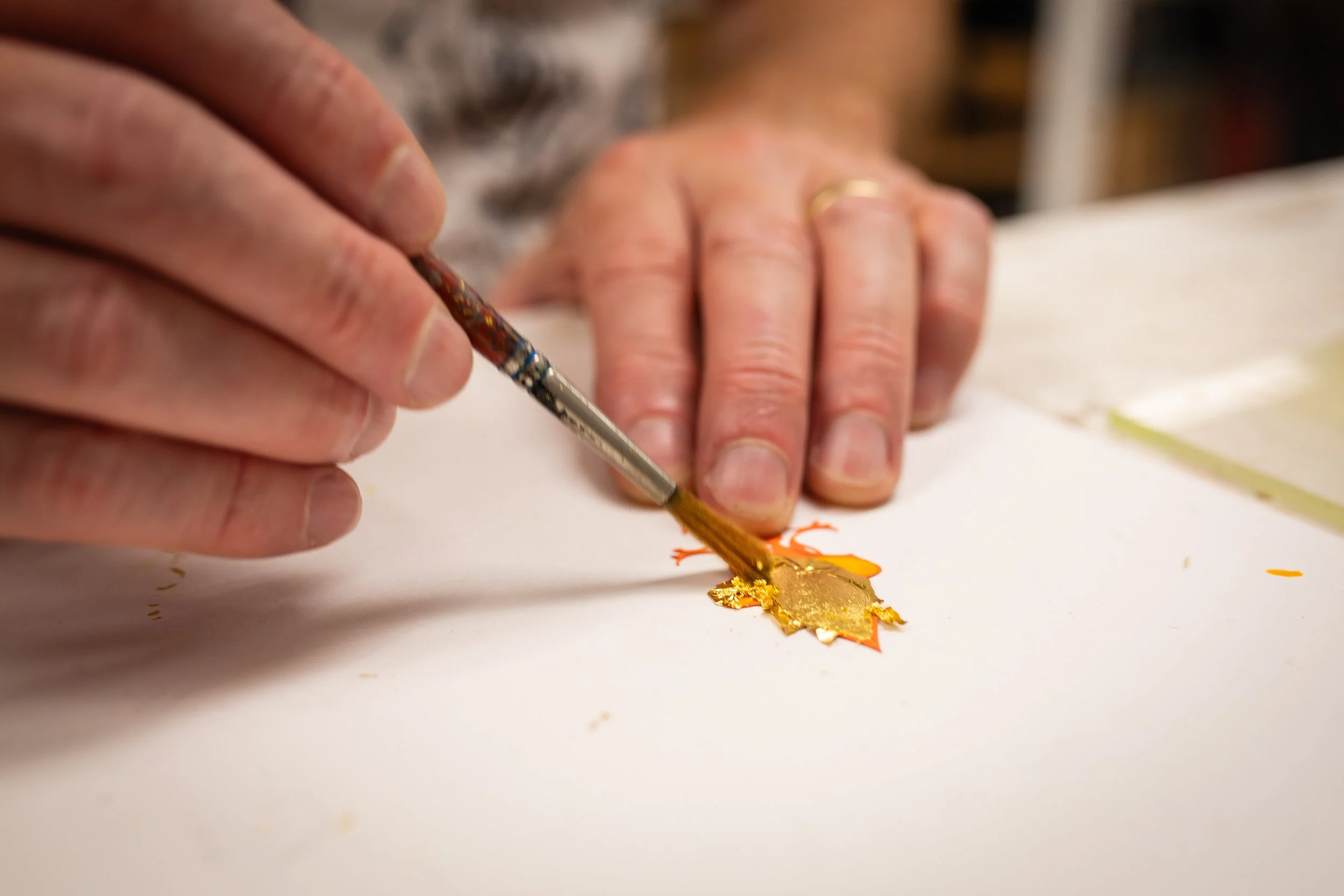Person painting a gold and orange leaf on white paper with a small paintbrush.