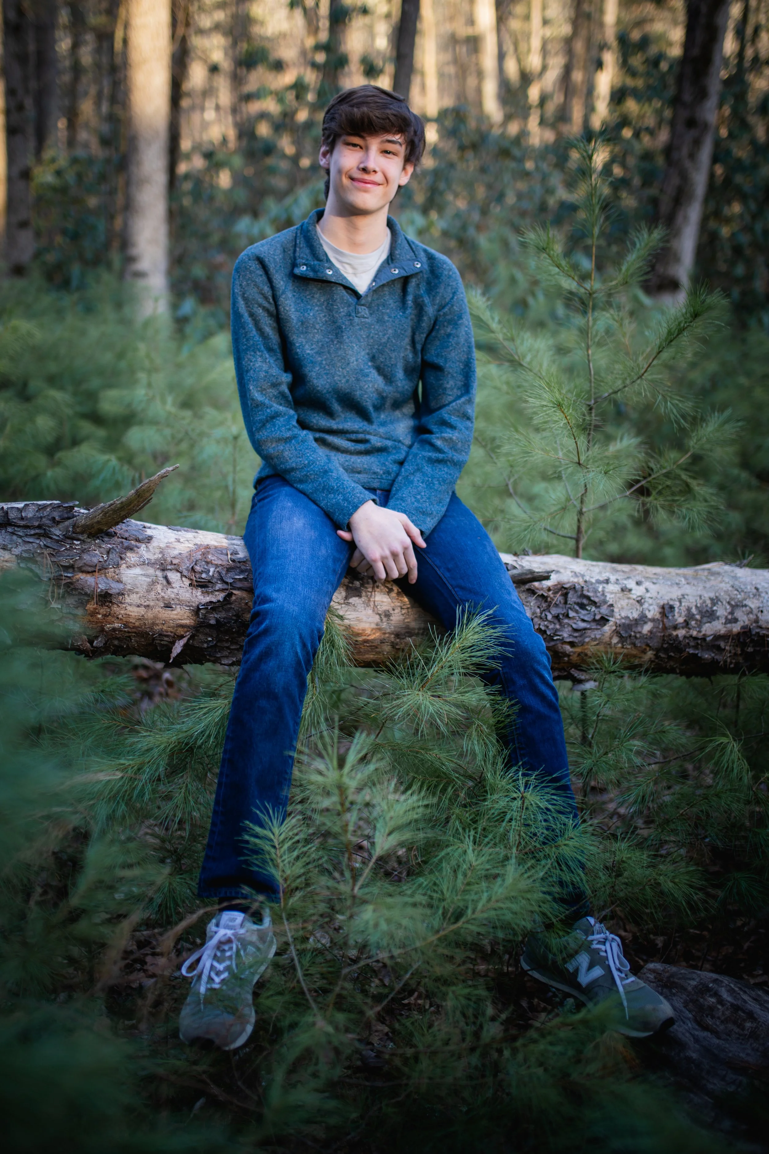 A young man sits on a fallen tree in a forest, smiling at the camera. He's wearing a gray sweater, blue jeans, and gray sneakers, surrounded by green pine trees.
