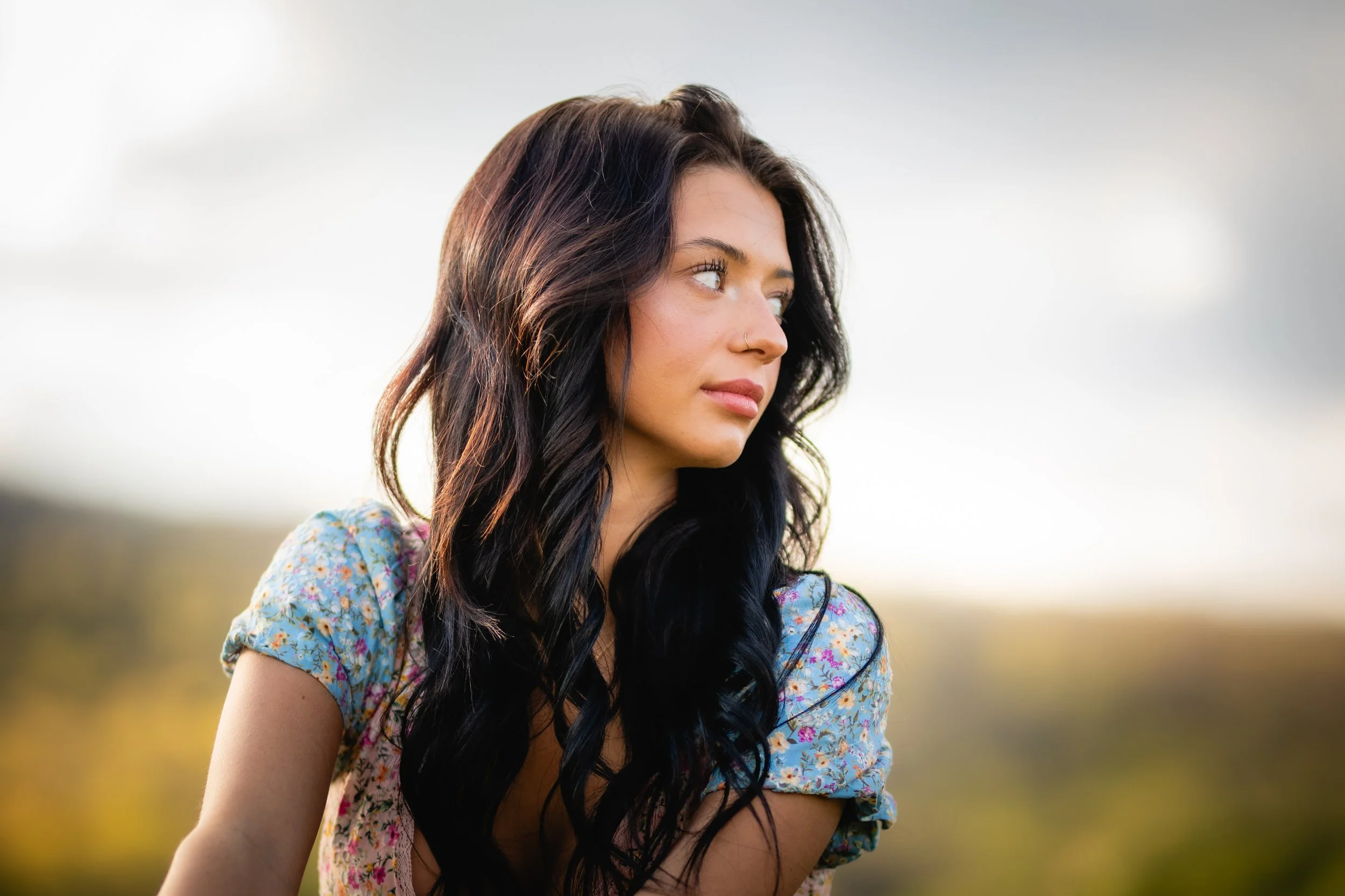 A young woman with long dark hair and a nose piercing looking to her right outdoors with a blurred nature background.