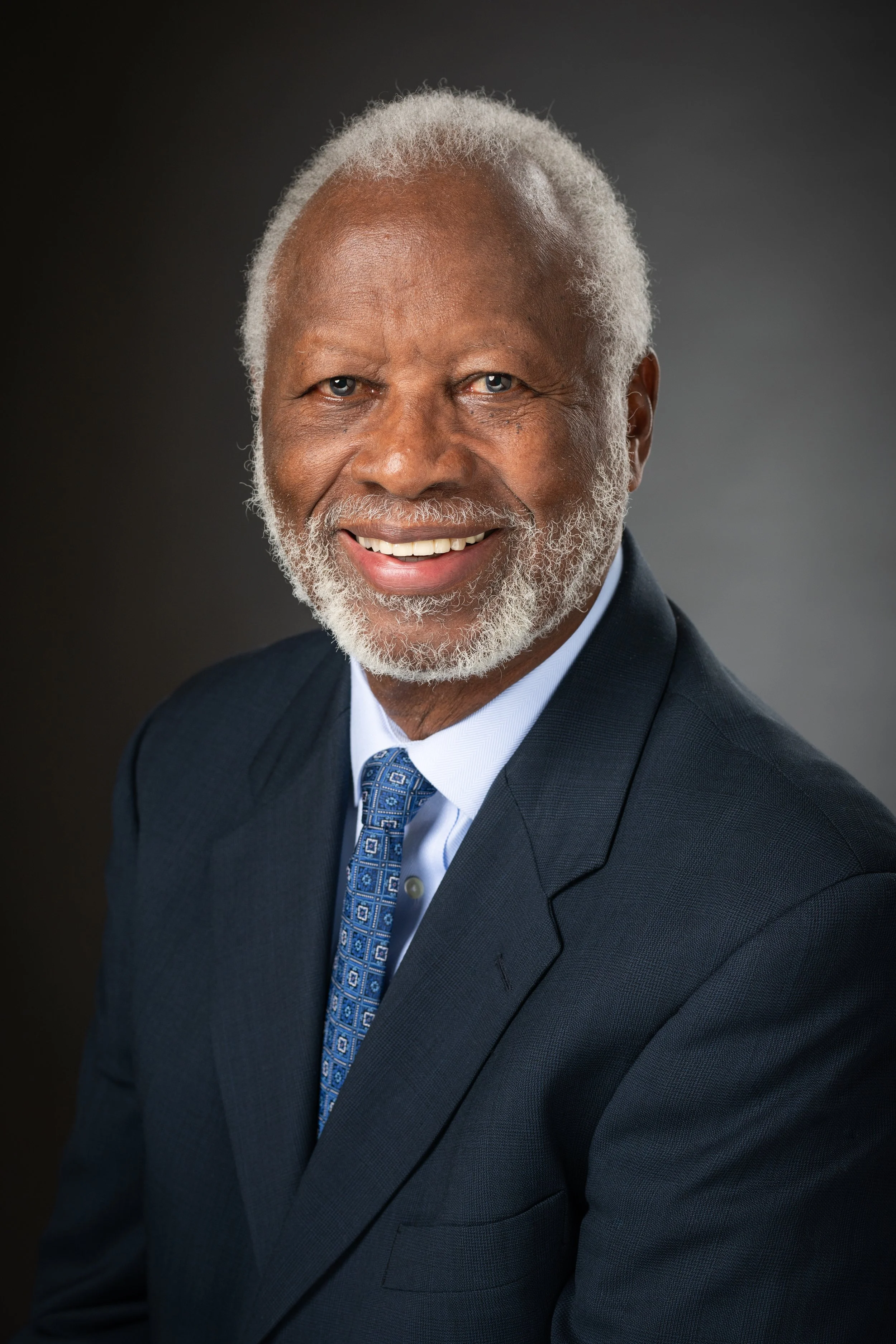 A smiling elderly African American man with gray hair and a beard, wearing a dark suit, light blue shirt, and patterned tie, against a dark background.