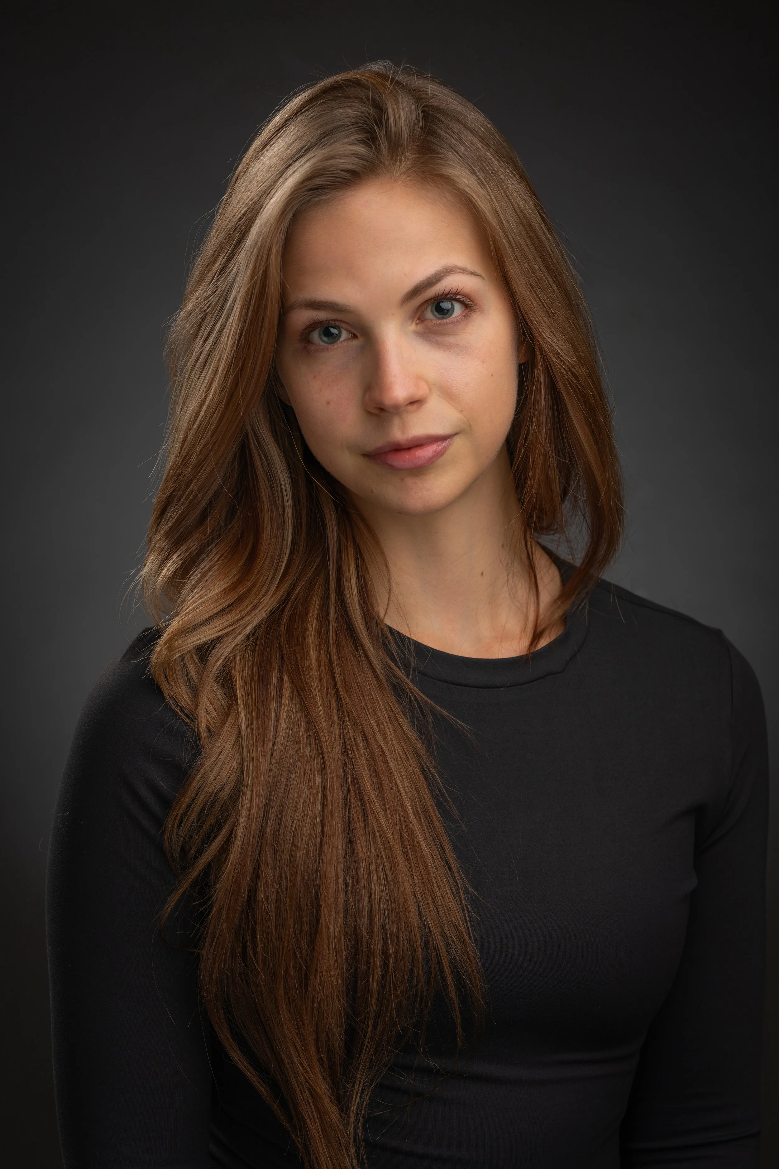 A woman with long, light brown hair and blue eyes wearing a black top against a dark gray background.
