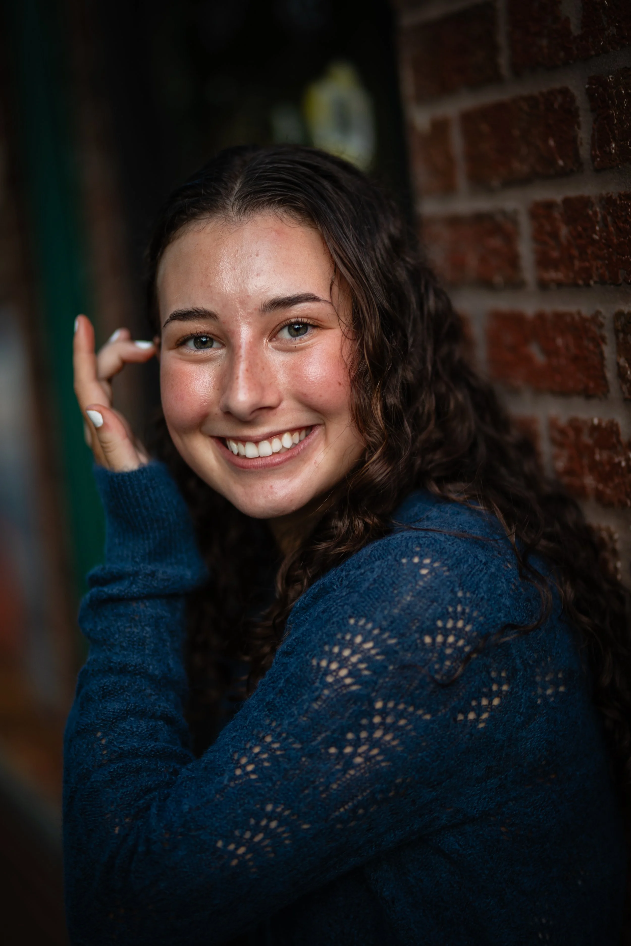 A young woman with curly brown hair and a bright smile, wearing a blue knitted sweater, standing against a brick wall.