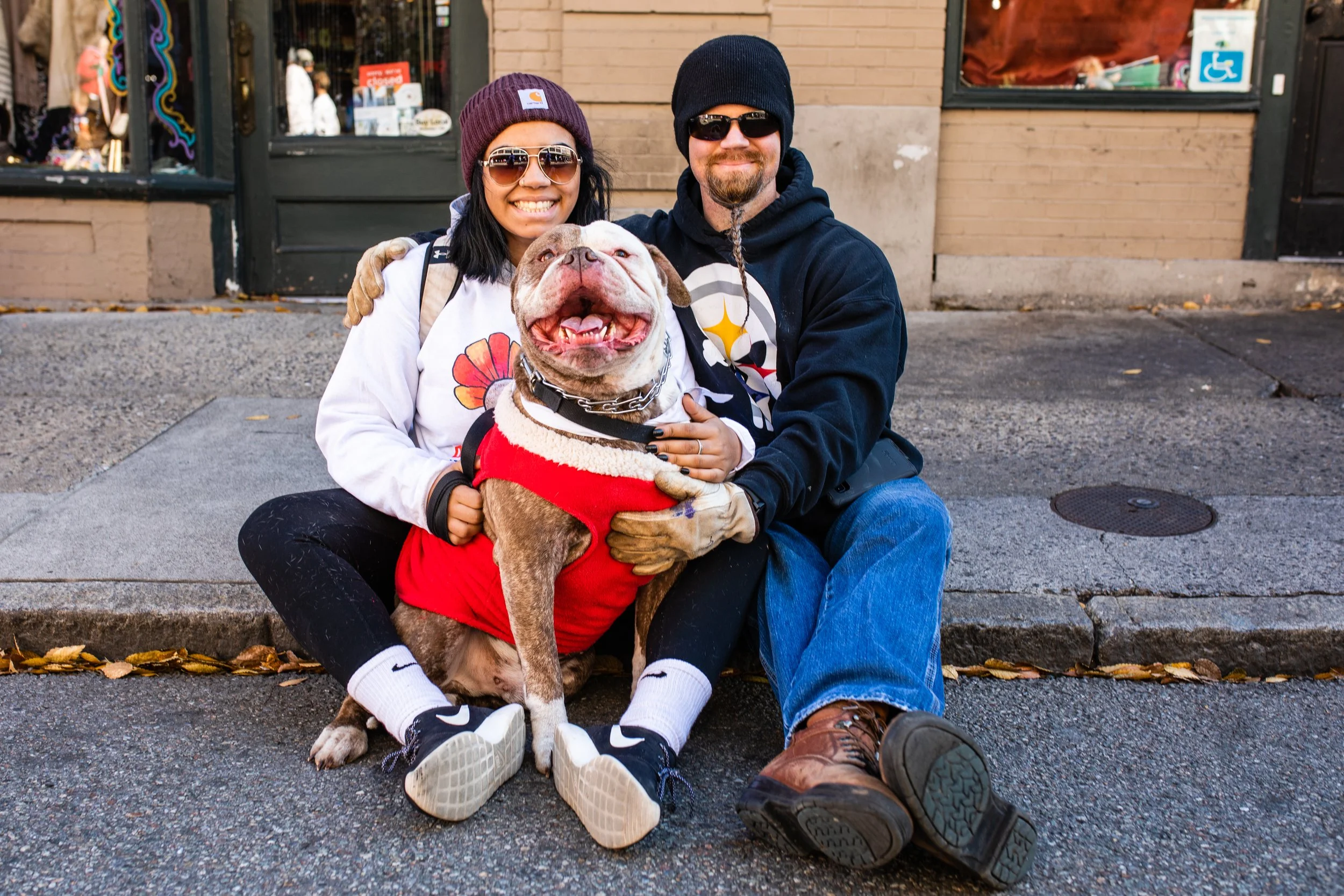 A smiling couple sitting on a sidewalk with a happy bulldog wearing a red sweater. The woman is wearing sunglasses and a beanie, while the man is wearing sunglasses, a beanie, and a hoodie. They are outdoors on a city street.