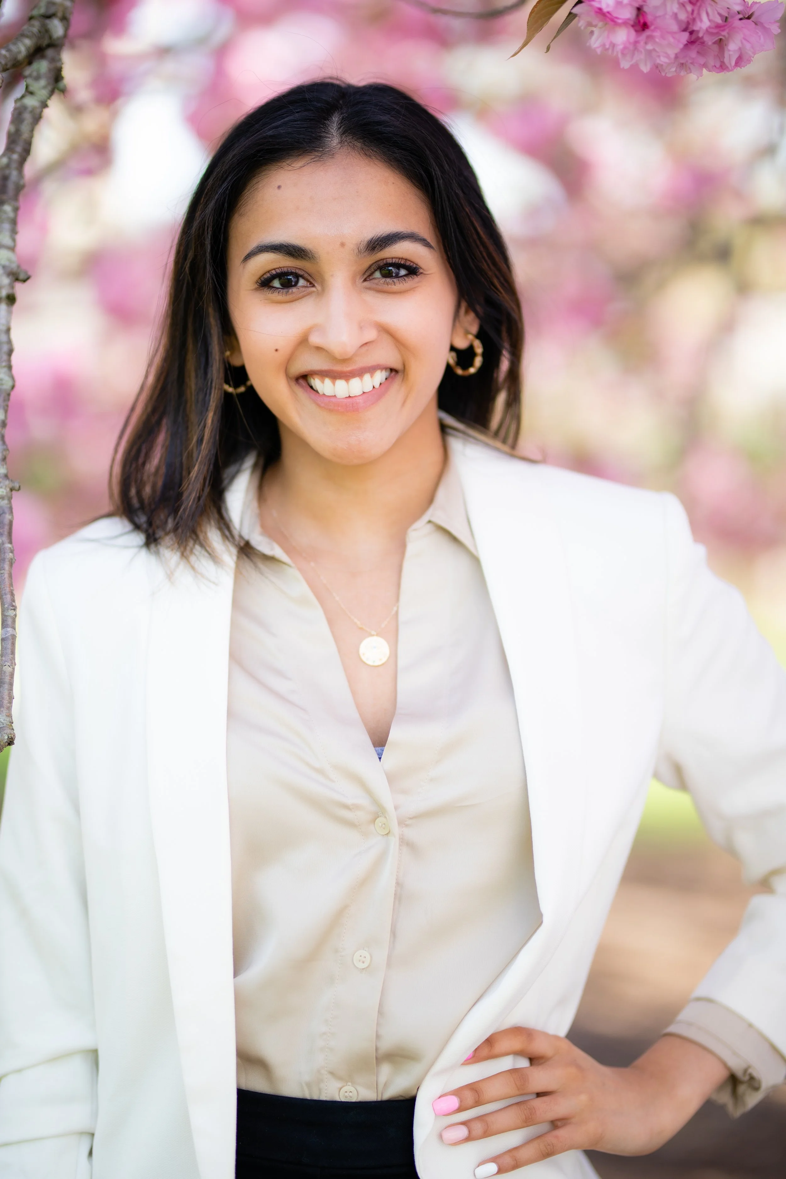 A woman smiling outdoors in front of pink flowering trees, wearing a white blazer and beige blouse.