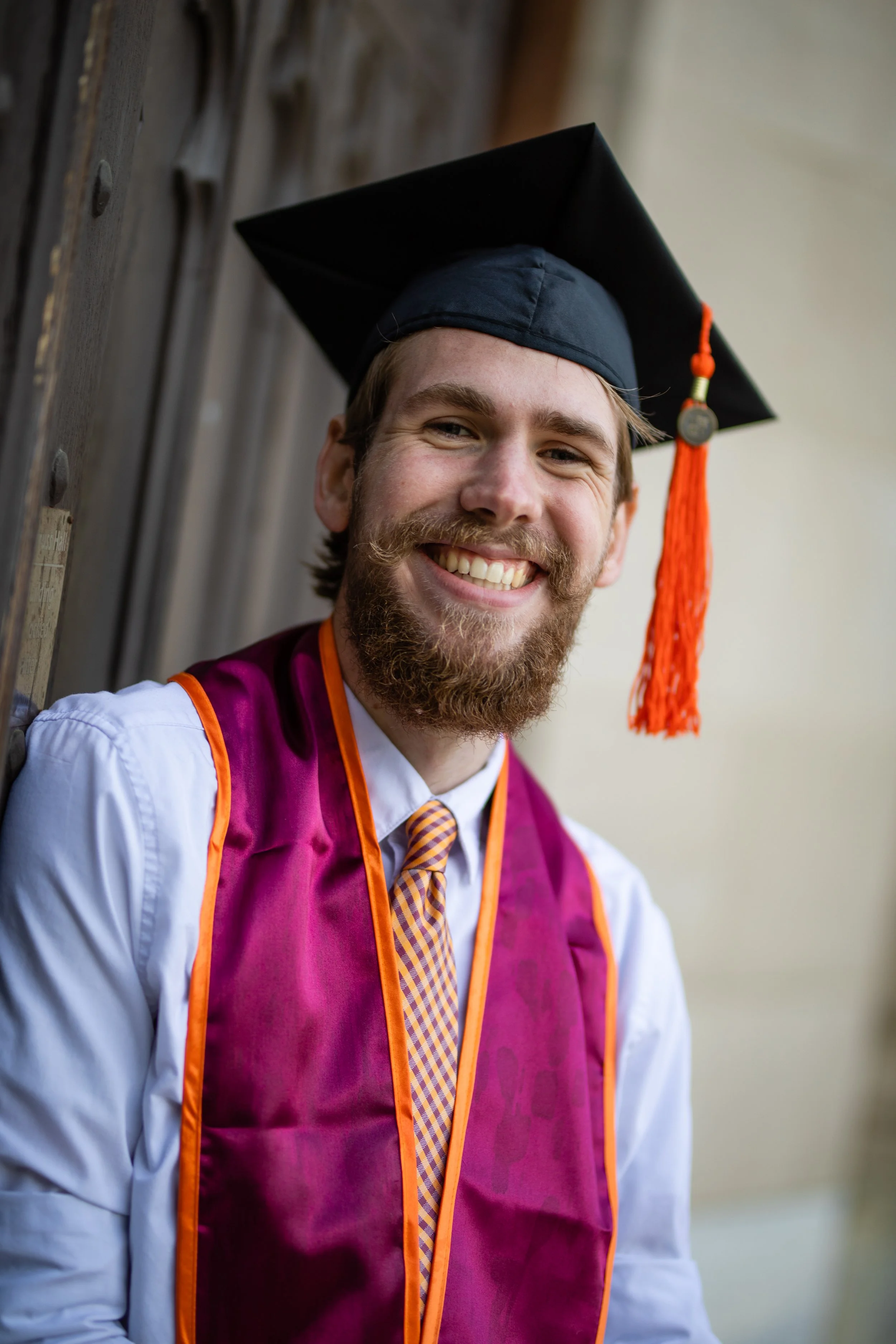 Smiling man wearing graduation cap and gown, standing outdoors against wooden background.