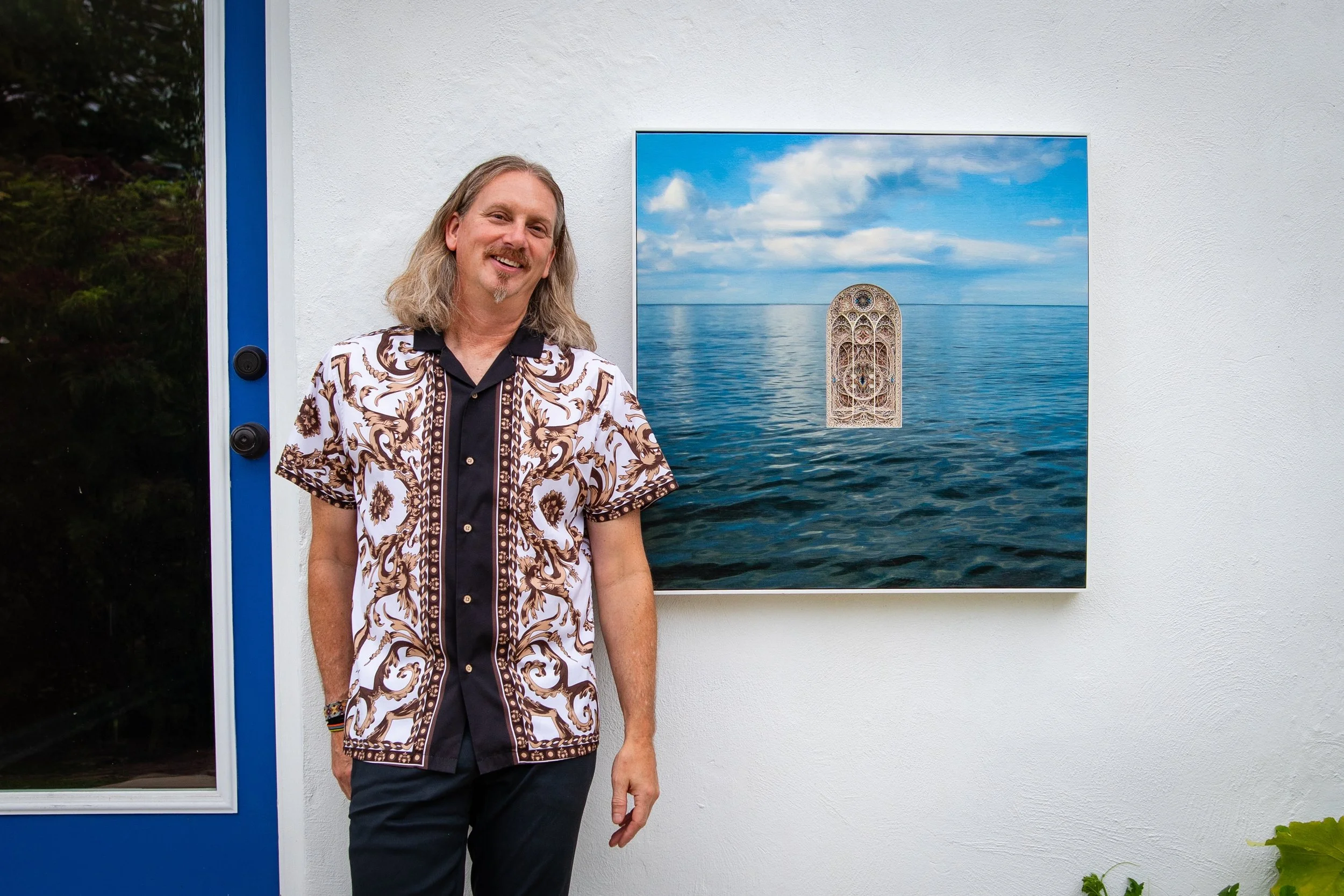 A man with long blond hair and a mustache, wearing a patterned short-sleeve shirt, standing next to a large photograph of a wispy sky, water, and a stained glass window inset, displayed on a white wall outside.