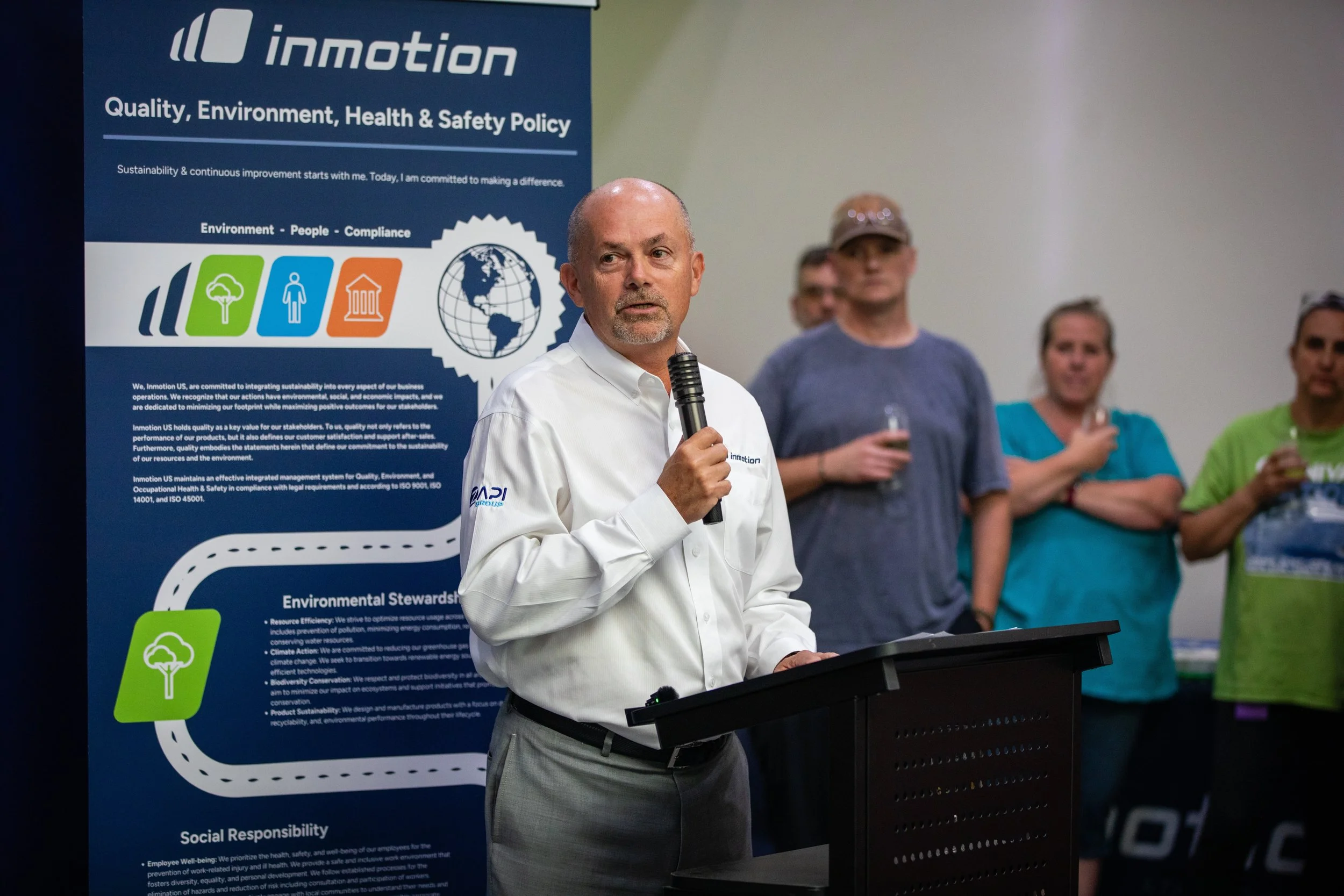 A man in a white shirt speaking into a microphone at a podium, with a blue environmental policy poster behind him and people in casual clothing holding drinks in the background.