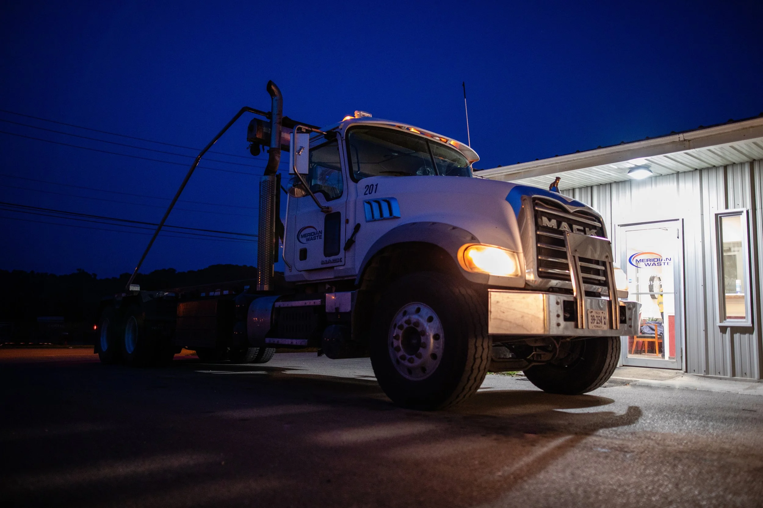 A white tow truck parked outside a building at night, with its headlights on and a sign reading 'Meridian Waste' on the door.