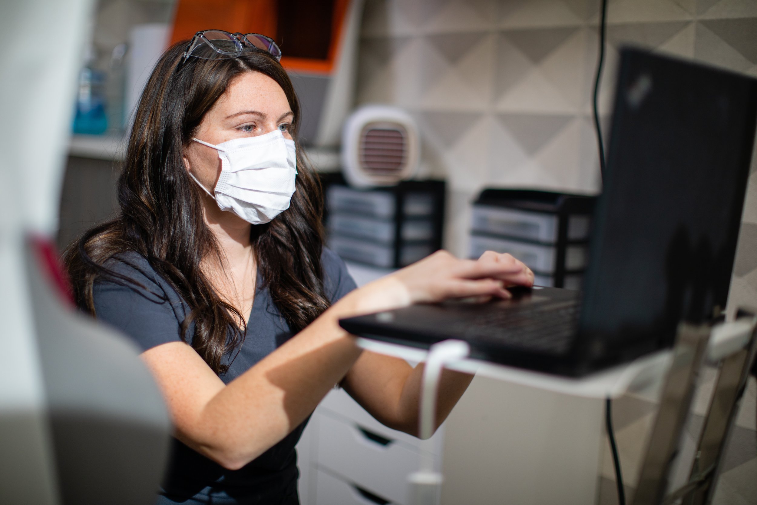 A woman with long brown hair wearing glasses and a face mask working on a laptop in a medical or laboratory setting.