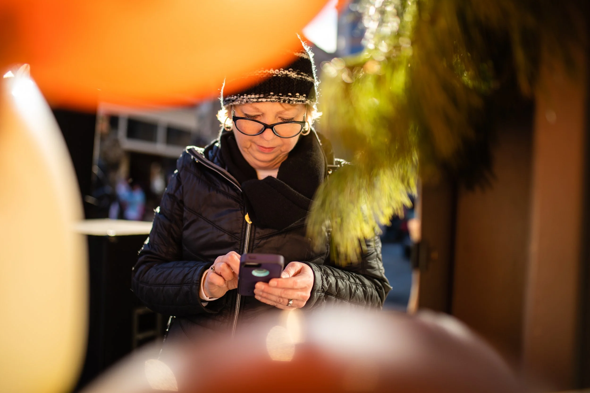 A woman with glasses and a black beanie looking at her phone, framed by colorful balloons.