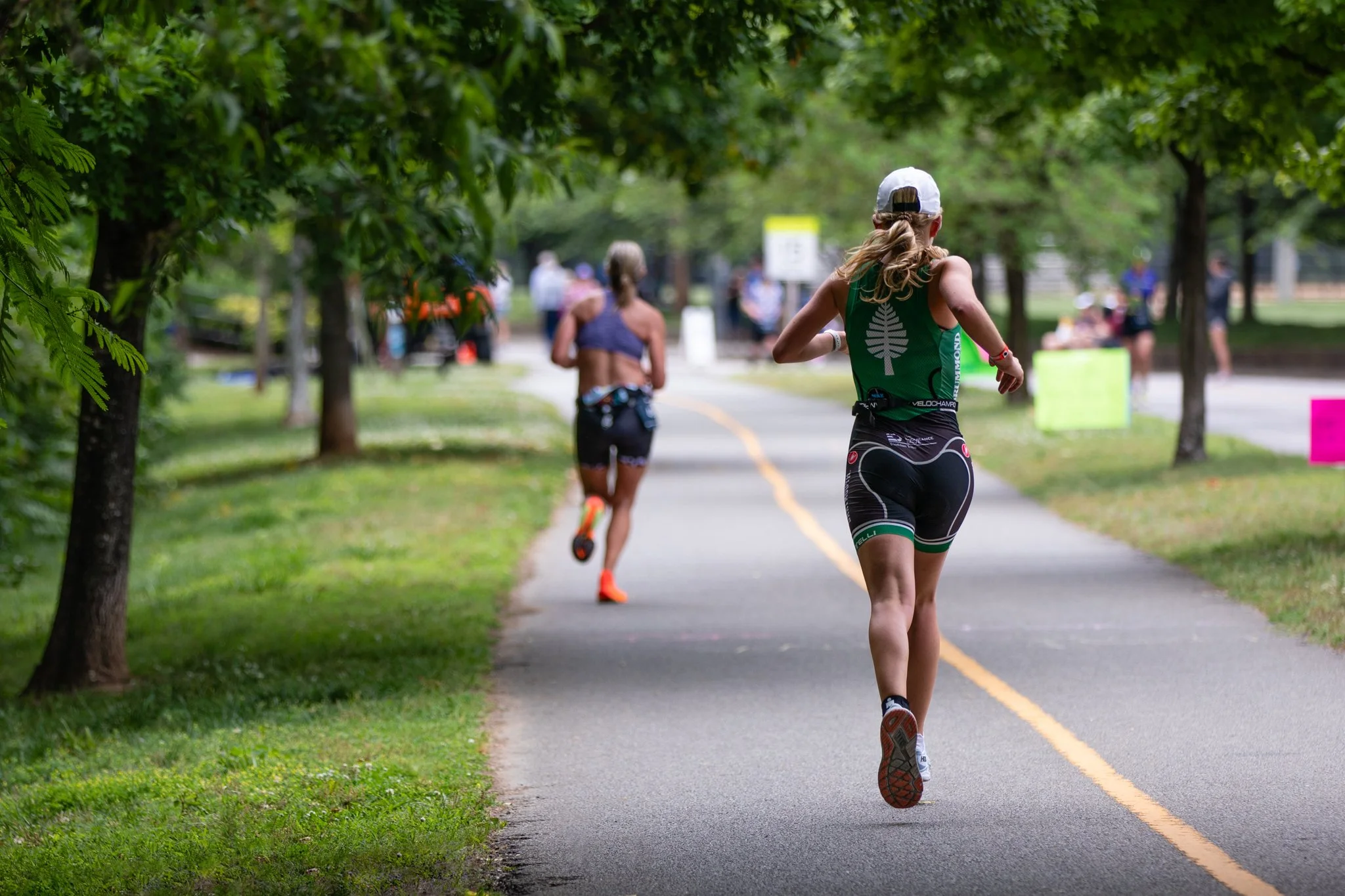 Two women running on a tree-lined paved path in a park, with several people in the background and some colorful signs.