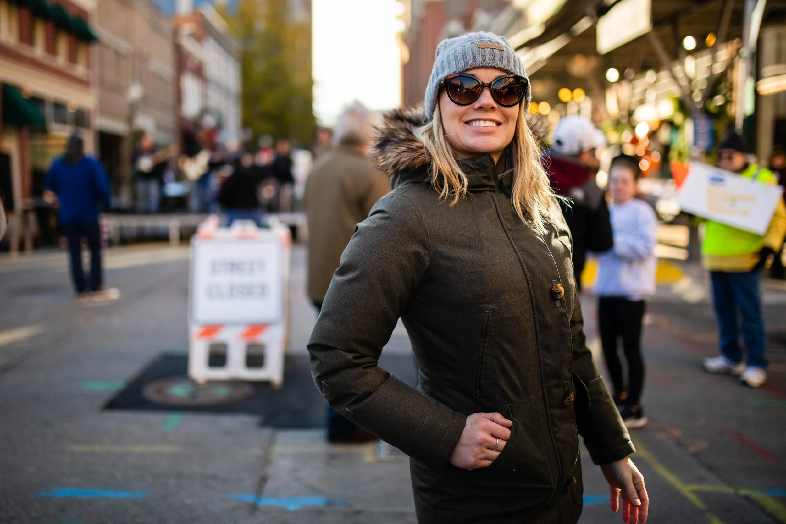 A woman wearing sunglasses, a gray beanie, and a black winter coat standing outdoors on a city street with a smile, with people and a 'Street Closed' sign in the background.