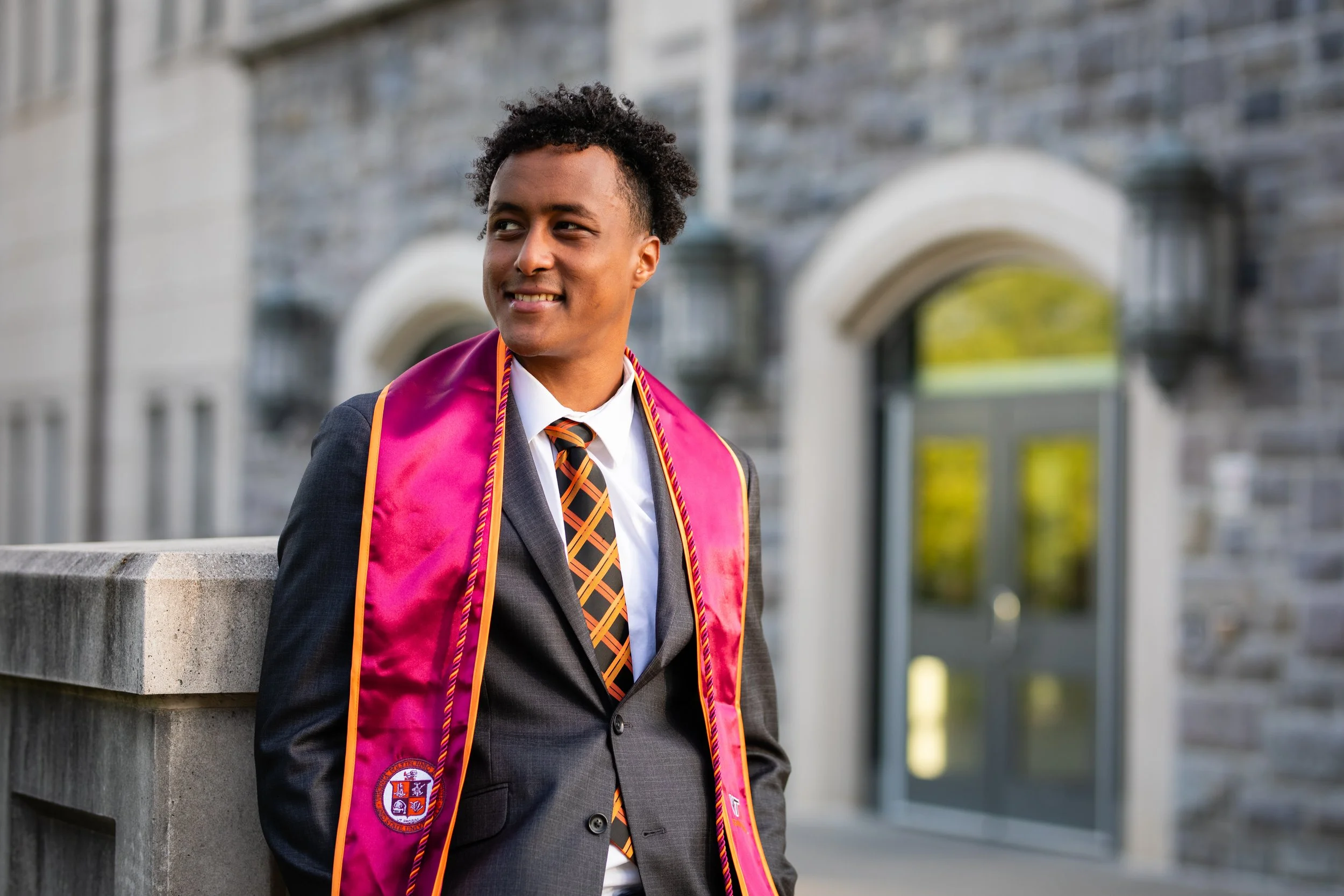 A young man in a suit and tie, wearing a pink graduation stole, stands outside near a stone building with arched windows, smiling and looking to the side.