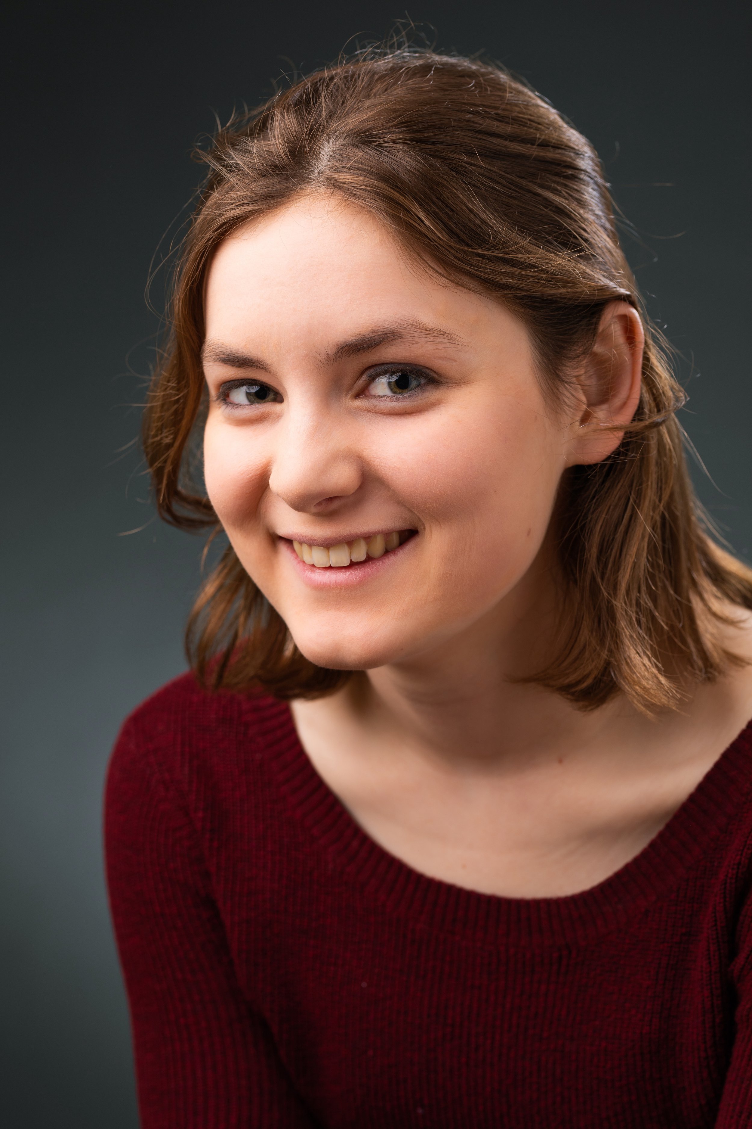 A young woman with short brown hair, light skin, and blue eyes smiling at the camera. She is wearing a maroon sweater and has a neutral gray background.