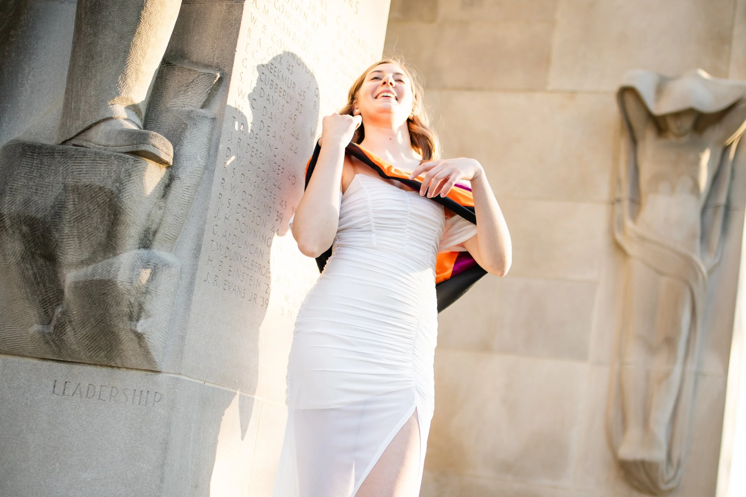 A young woman in a white dress standing outdoors, smiling, with a colorful sash over her shoulder, near a stone monument with carved inscriptions and sculptures.