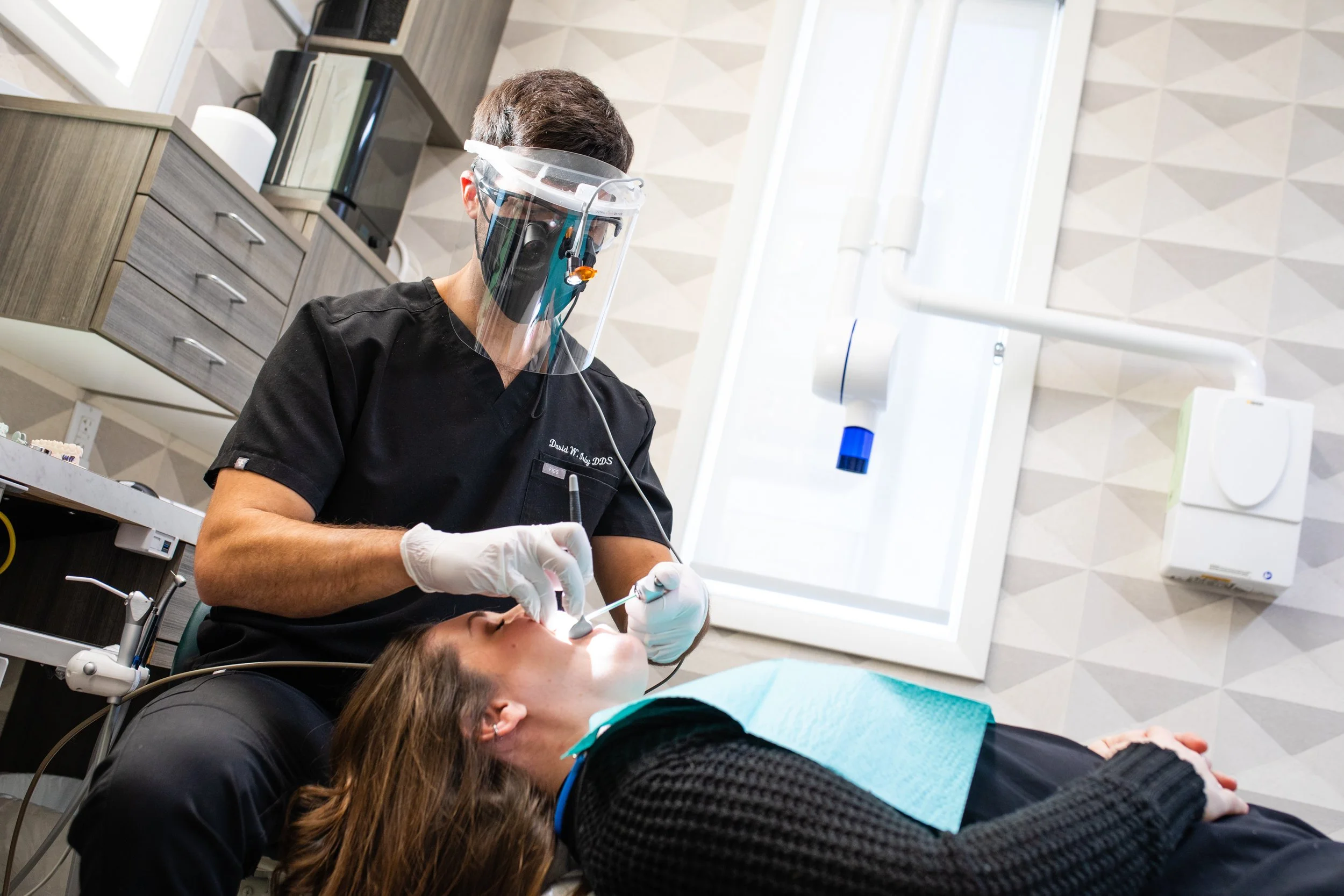 Dentist performing a dental procedure on a patient lying on a dental chair, both wearing protective gear in a modern dental office.