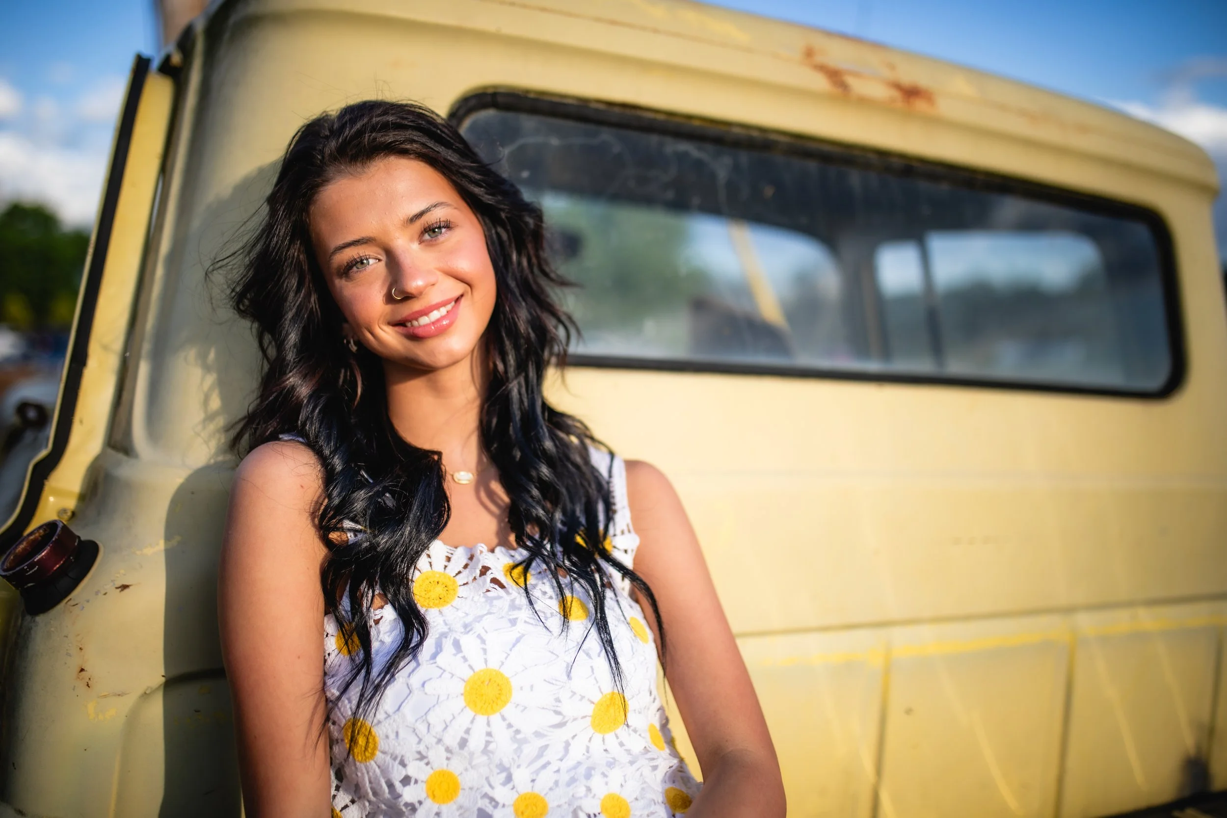 A young woman with dark curly hair, a nose ring, and a white sleeveless top with yellow daisies, smiling and leaning against a yellow vintage vehicle outdoors.
