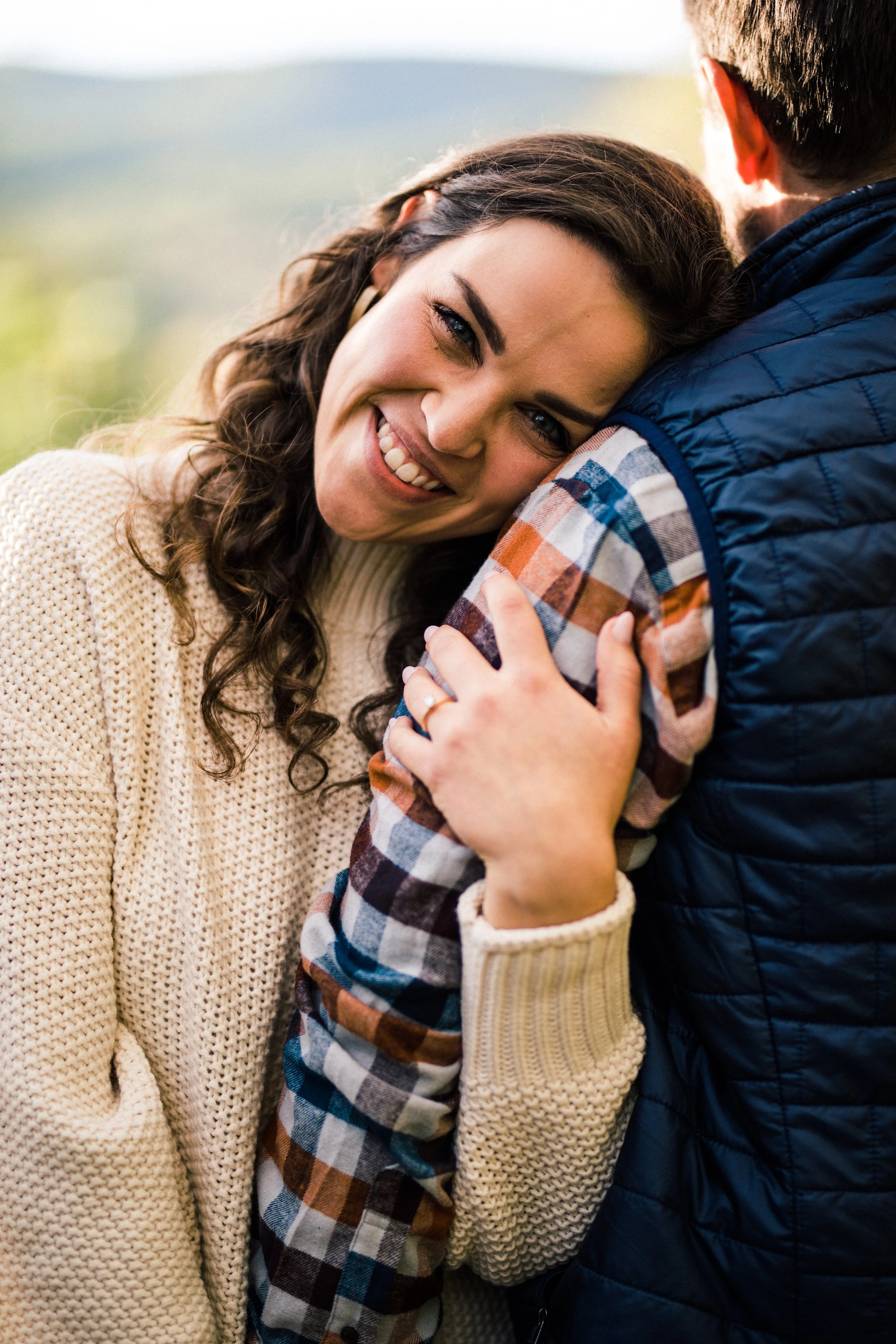 A woman with curly hair smiling and hugging a man, both outdoors on a bright day.
