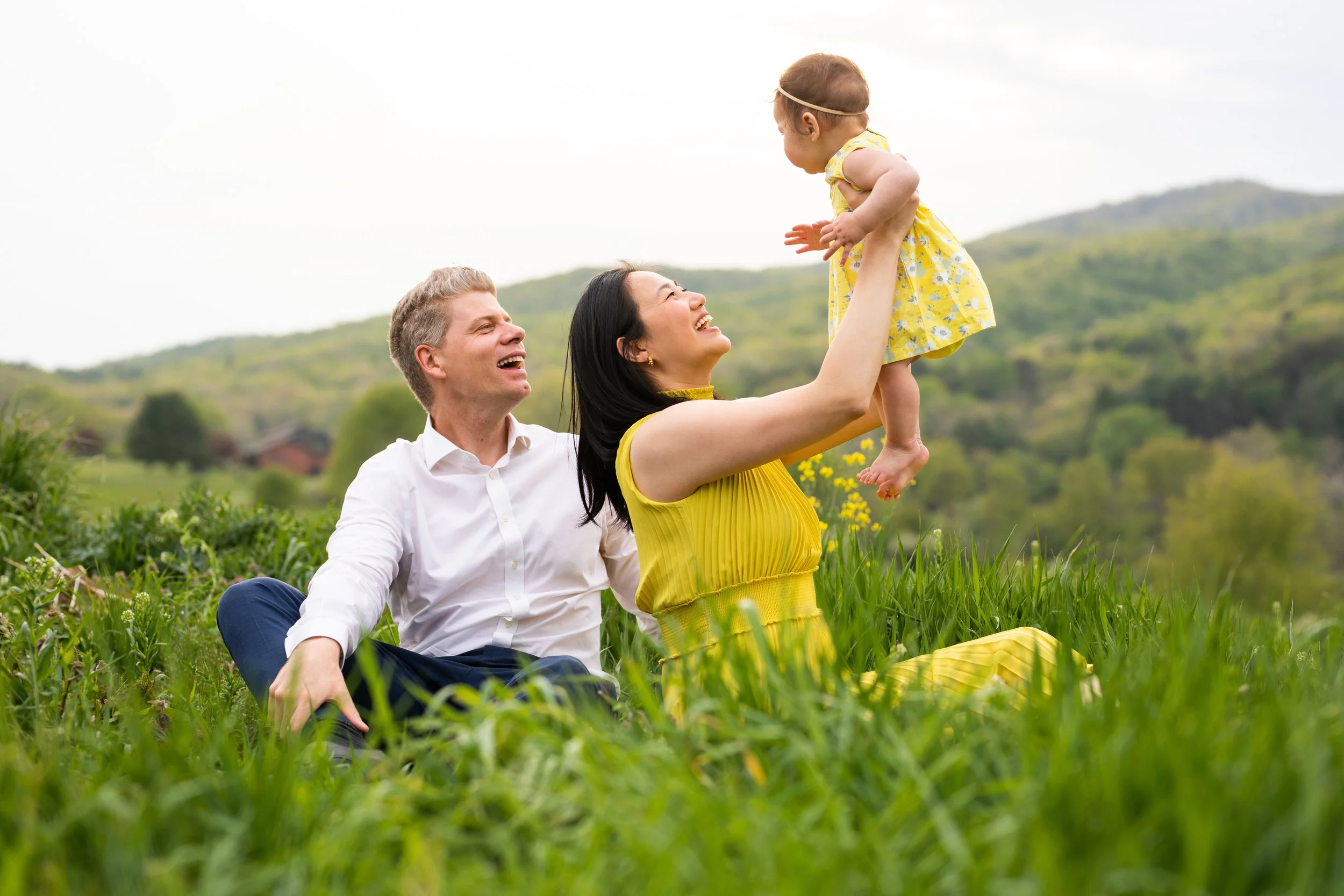 A family of three - a man, a woman, and a toddler girl - enjoying time outdoors in a grassy field, with the man sitting on the grass, the woman holding the girl up in the air, all smiling happily against a backdrop of green hills.