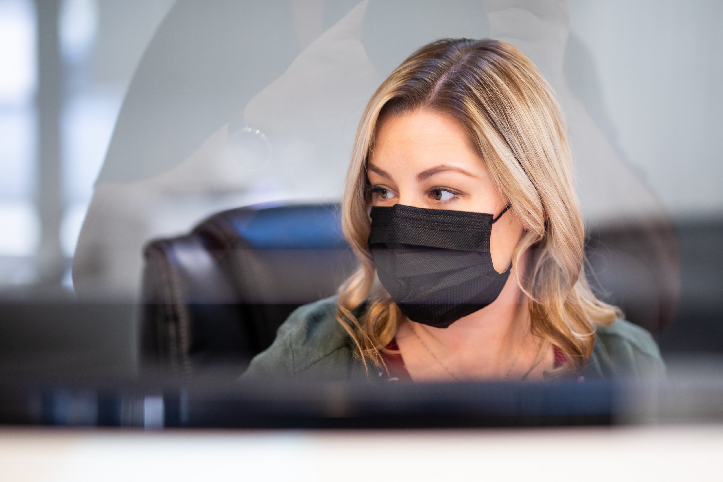 A woman with blonde hair wearing a black face mask, looking to her right inside an office, with a glass partition in front of her.