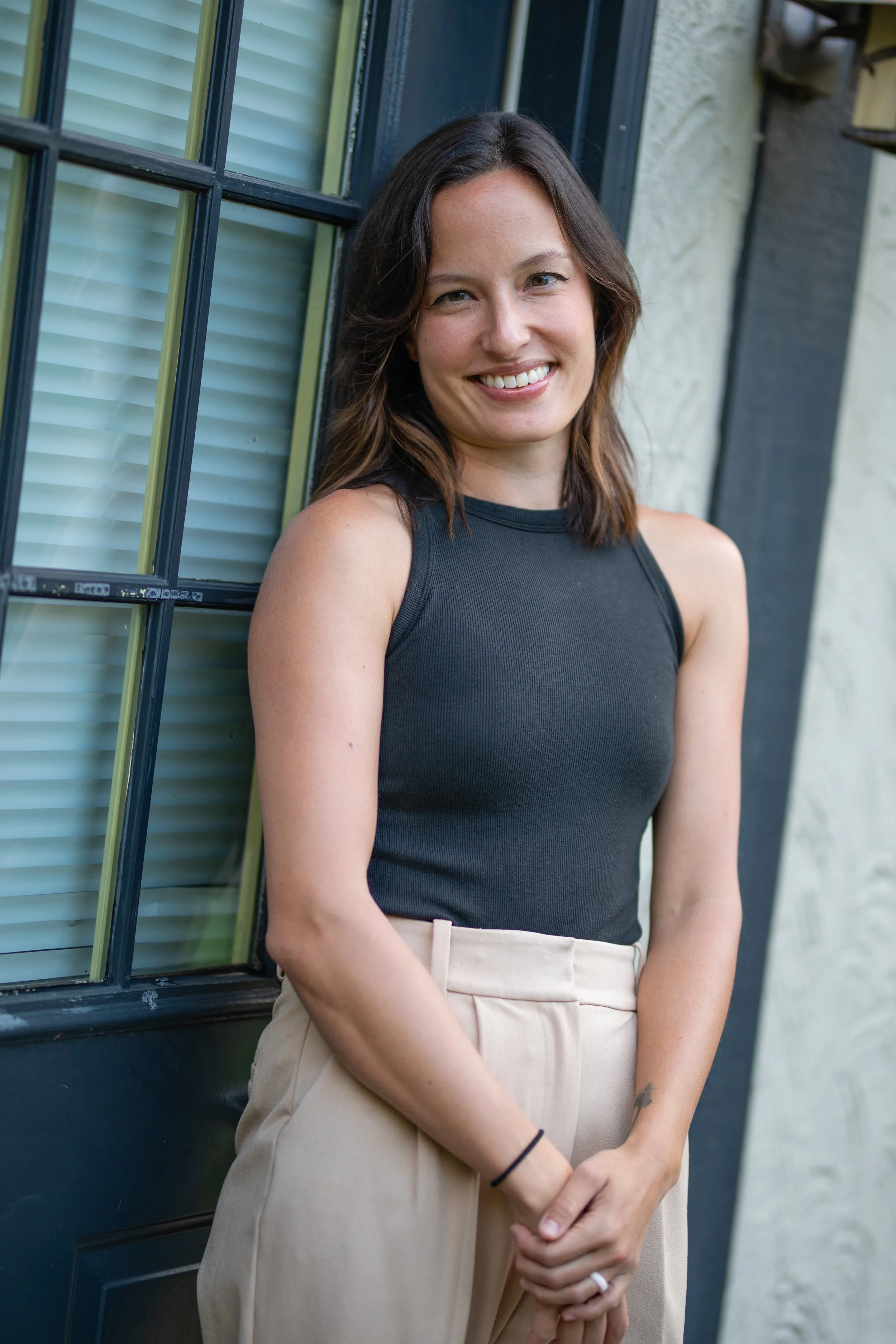 Smiling woman with shoulder-length brown hair wearing a black sleeveless top and beige pants standing outdoors next to a window with blinds.