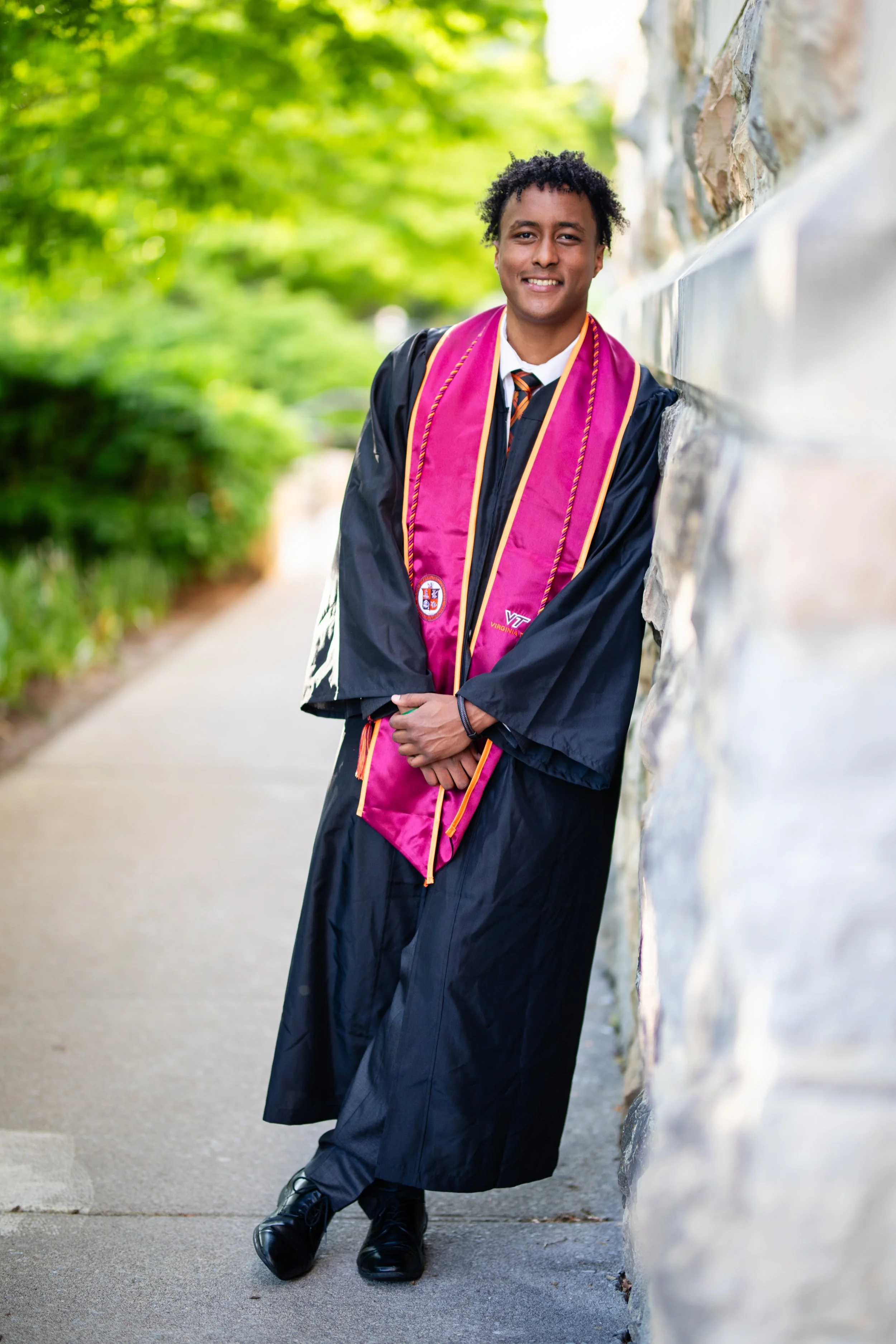 A young man in graduation cap and gown leaning against a stone wall, outdoors with green trees in the background.