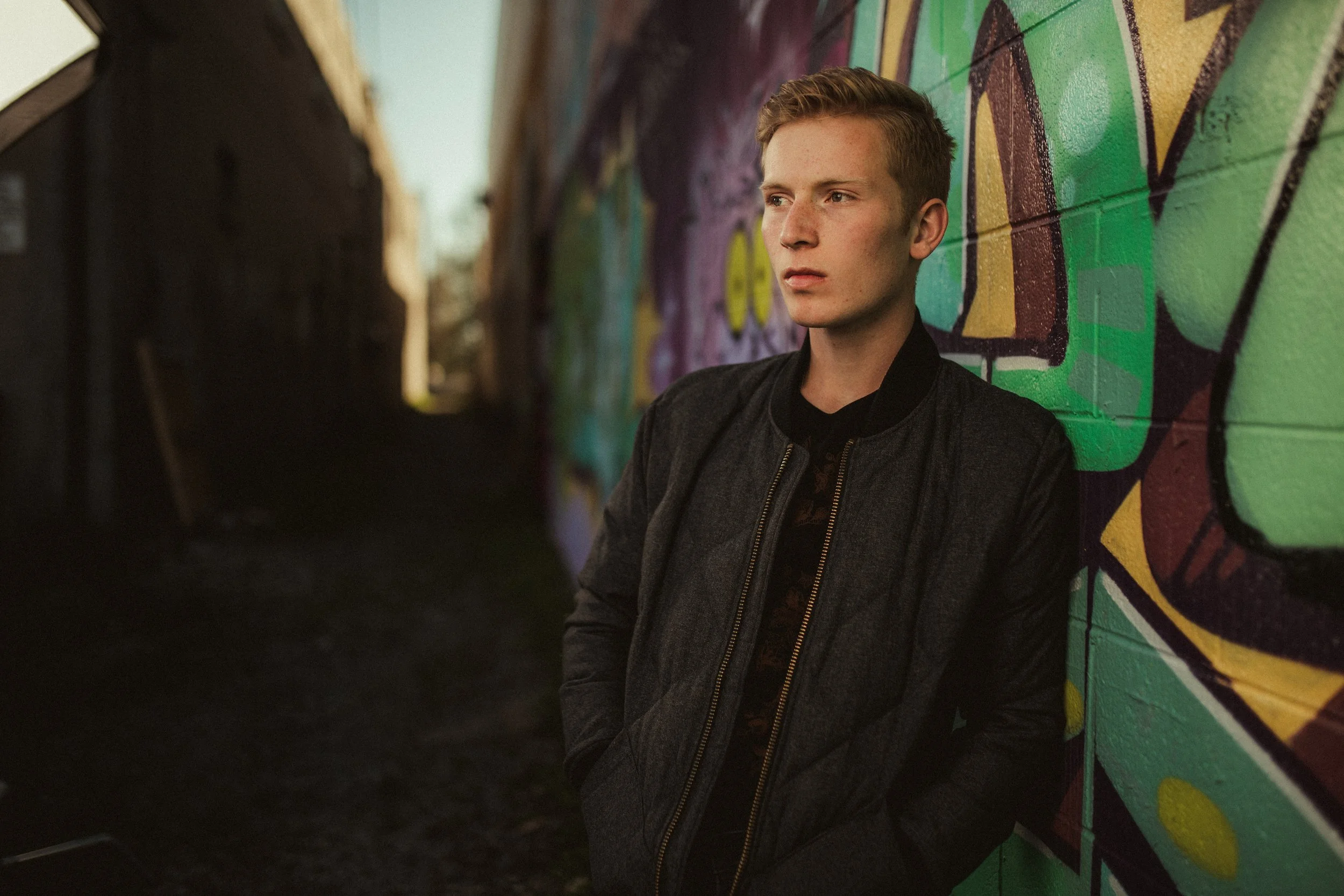 A young man with short blonde hair wearing a dark jacket leans against a graffiti-covered wall in an alleyway