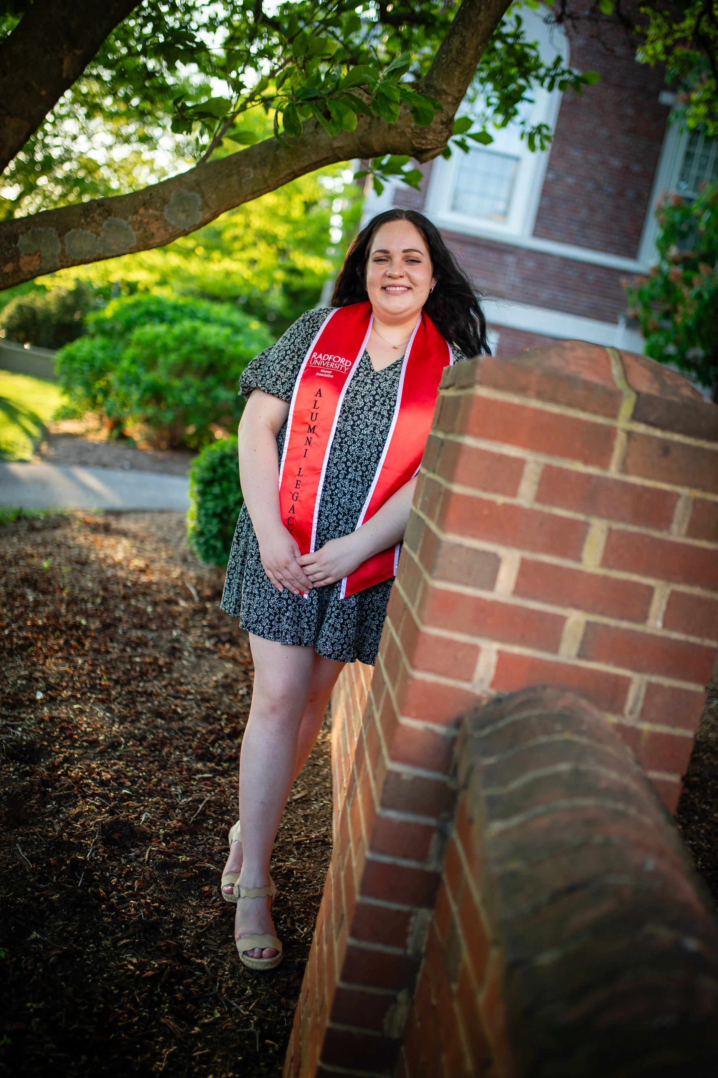 A young woman in a black patterned dress smiles and poses outdoors, wearing a red sash that reads 'Radford University Alumni Legacy,' standing next to a brick wall with green trees and a house in the background.