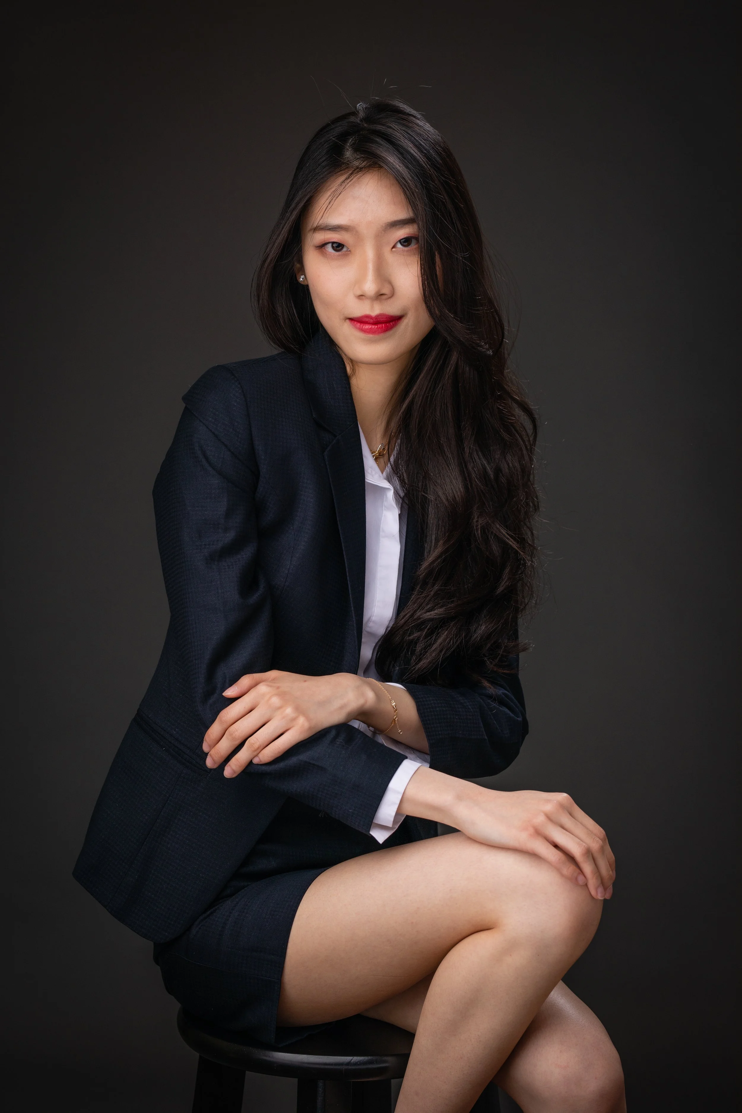 A woman with long dark hair sitting on a black stool against a dark grey background, wearing a dark blazer and skirt with a white shirt, looking at the camera with a slight smile.