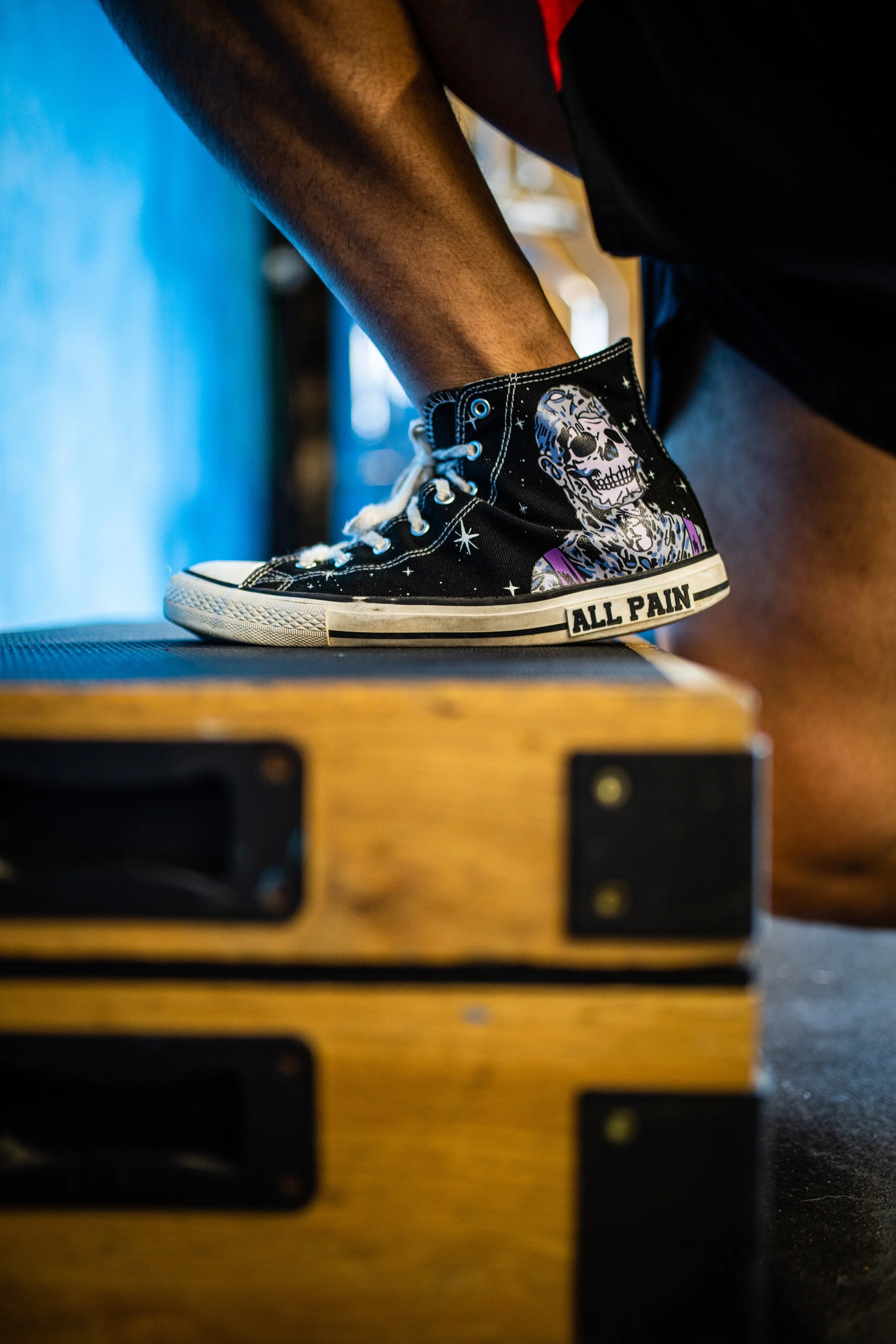 A person doing a box jump, wearing black sneakers with a colorful skeleton design and 'ALL PAIN' written on the sole.