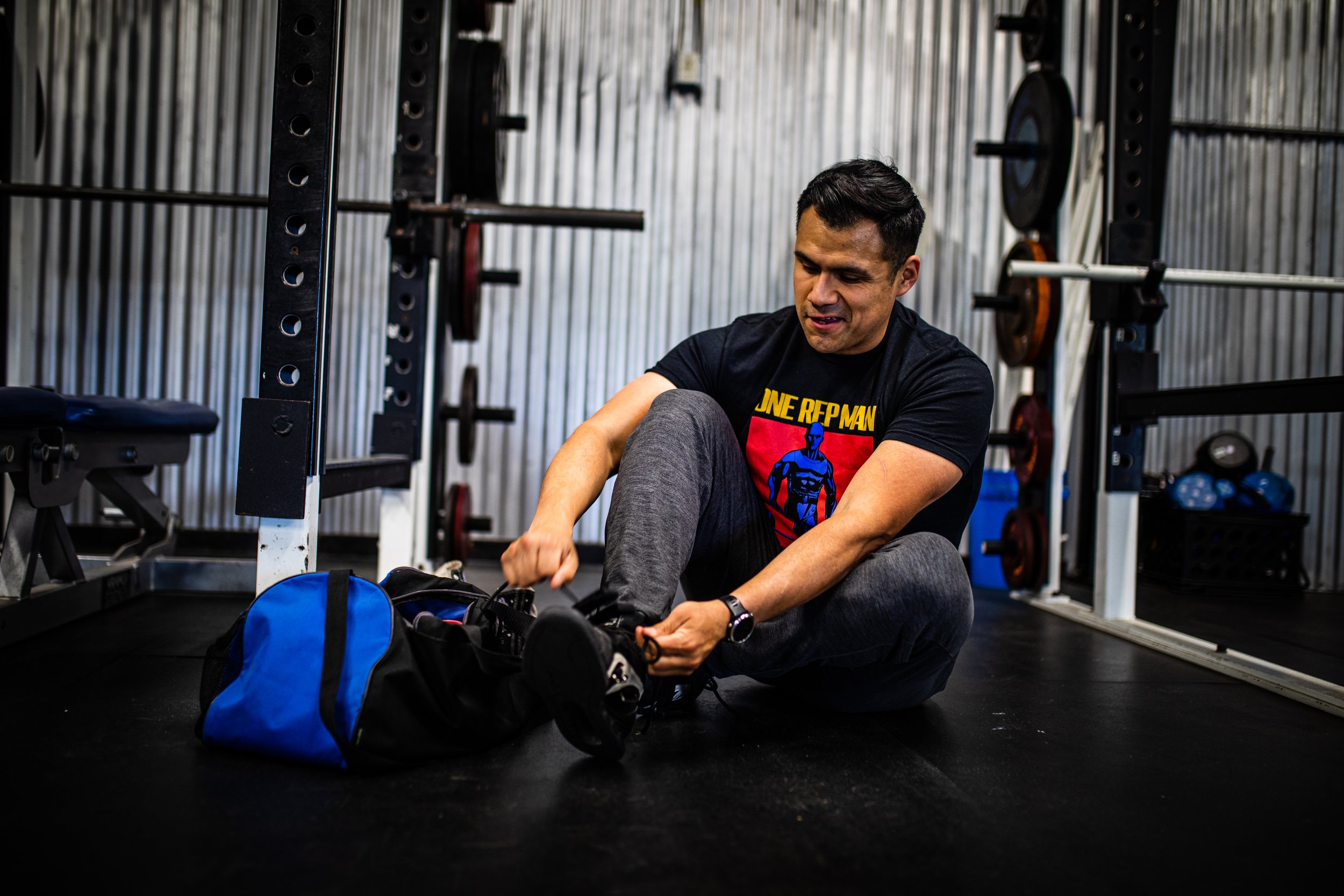 A man sitting on the gym floor, tying his shoelaces, with gym equipment and weights in the background.