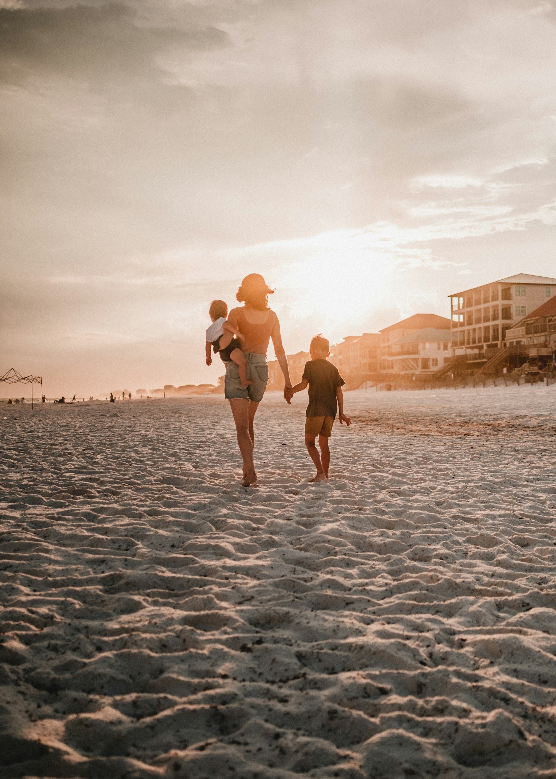 mother walking on the beach with her children; carrying one on her hip and holding another one's hand