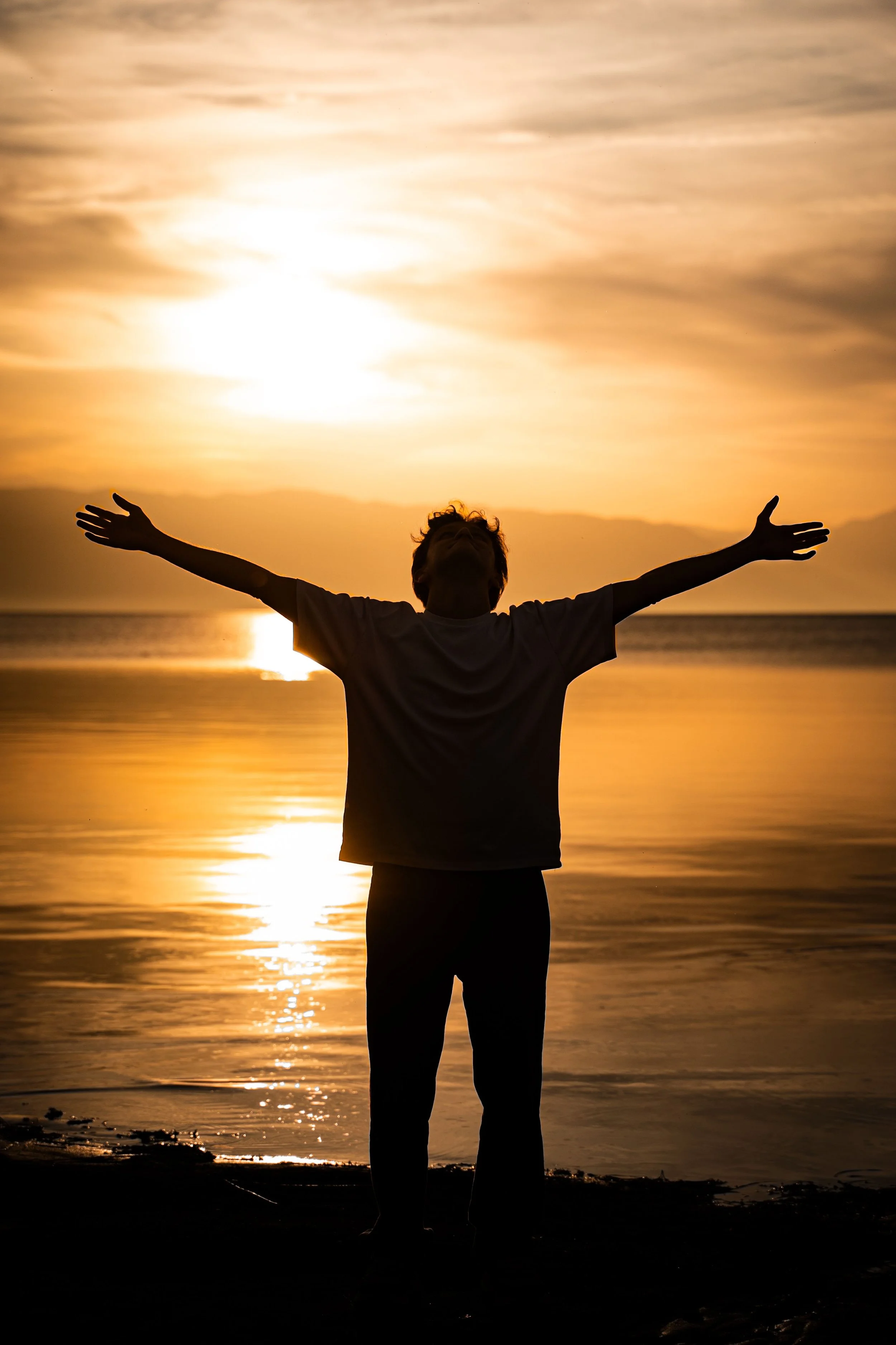 Person standing on the beach with arms outstretched during a golden sunset over the ocean.