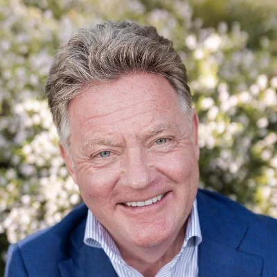A smiling middle-aged man with gray hair outdoors, wearing a blue blazer and white shirt, with blurred greenery in the background.