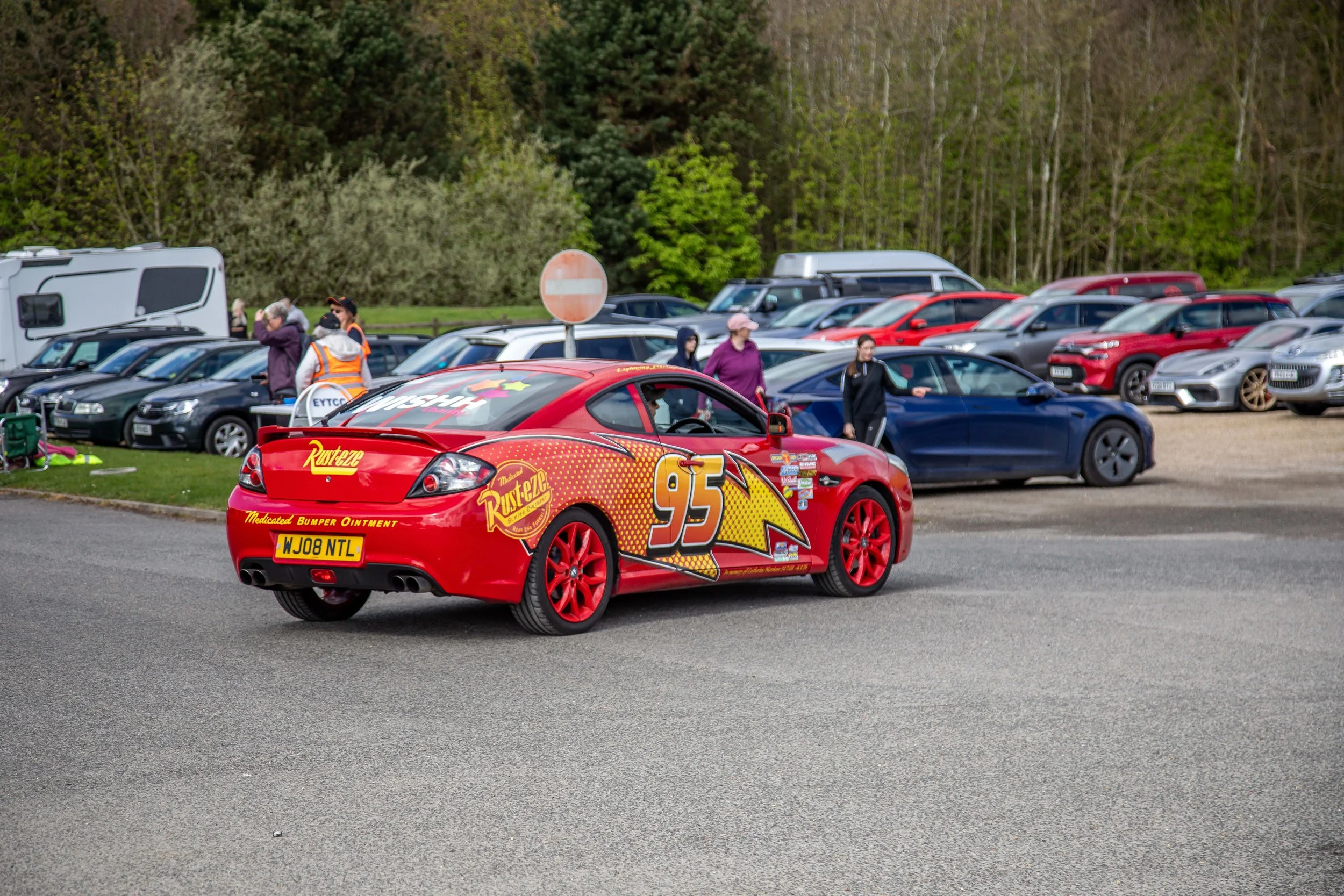 A red race car decorated with the number 95 and various sponsor logos, parked in a parking lot filled with other cars, with people standing nearby and trees in the background.